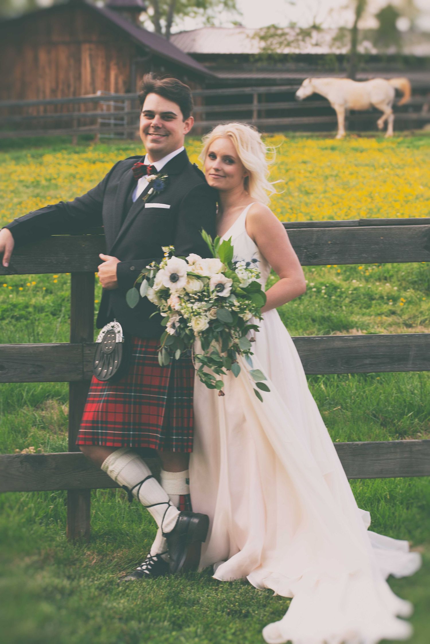 Bride and groom in Scottish kilt pose by wooden fence at farm wedding with white horse and yellow wildflowers in background.
