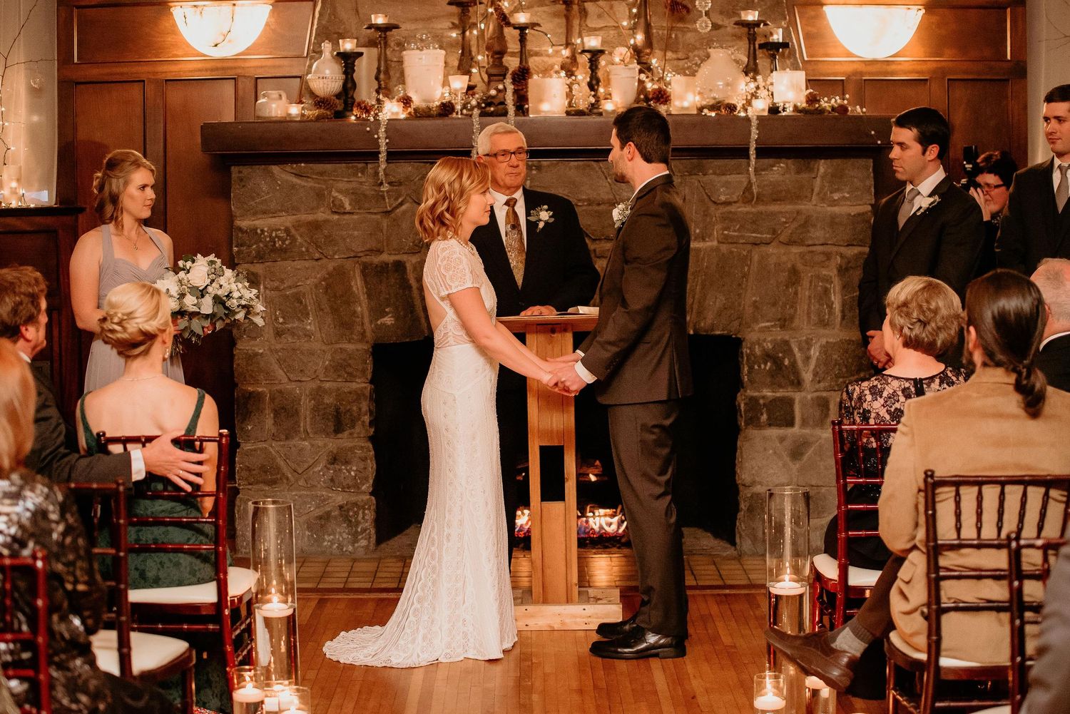 Couple exchanges vows at elegant indoor wedding ceremony with stone fireplace, candles, and guests seated in Chiavari chairs.