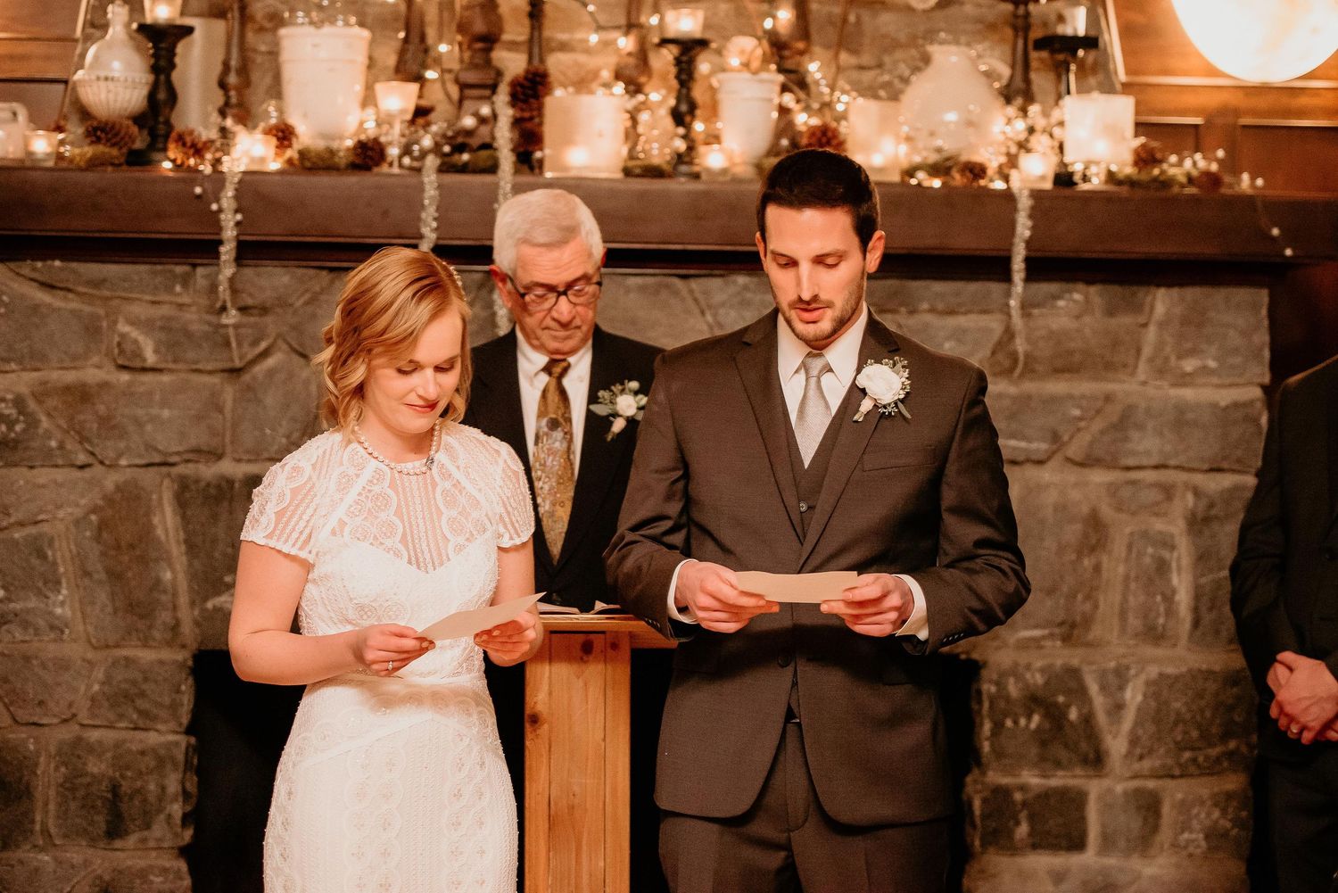 Bride and groom exchange vows at rustic wedding ceremony beside stone fireplace with warm candlelight.