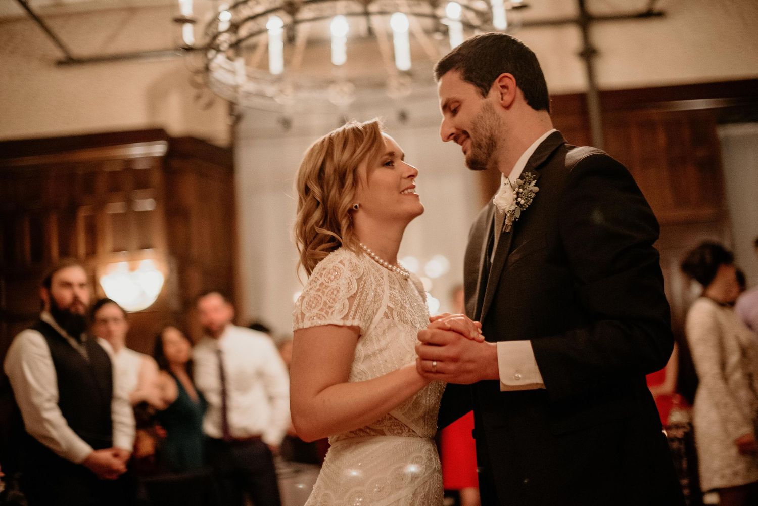 A bride and groom share their first dance, smiling at each other in an elegant warmly lit wedding reception venue.