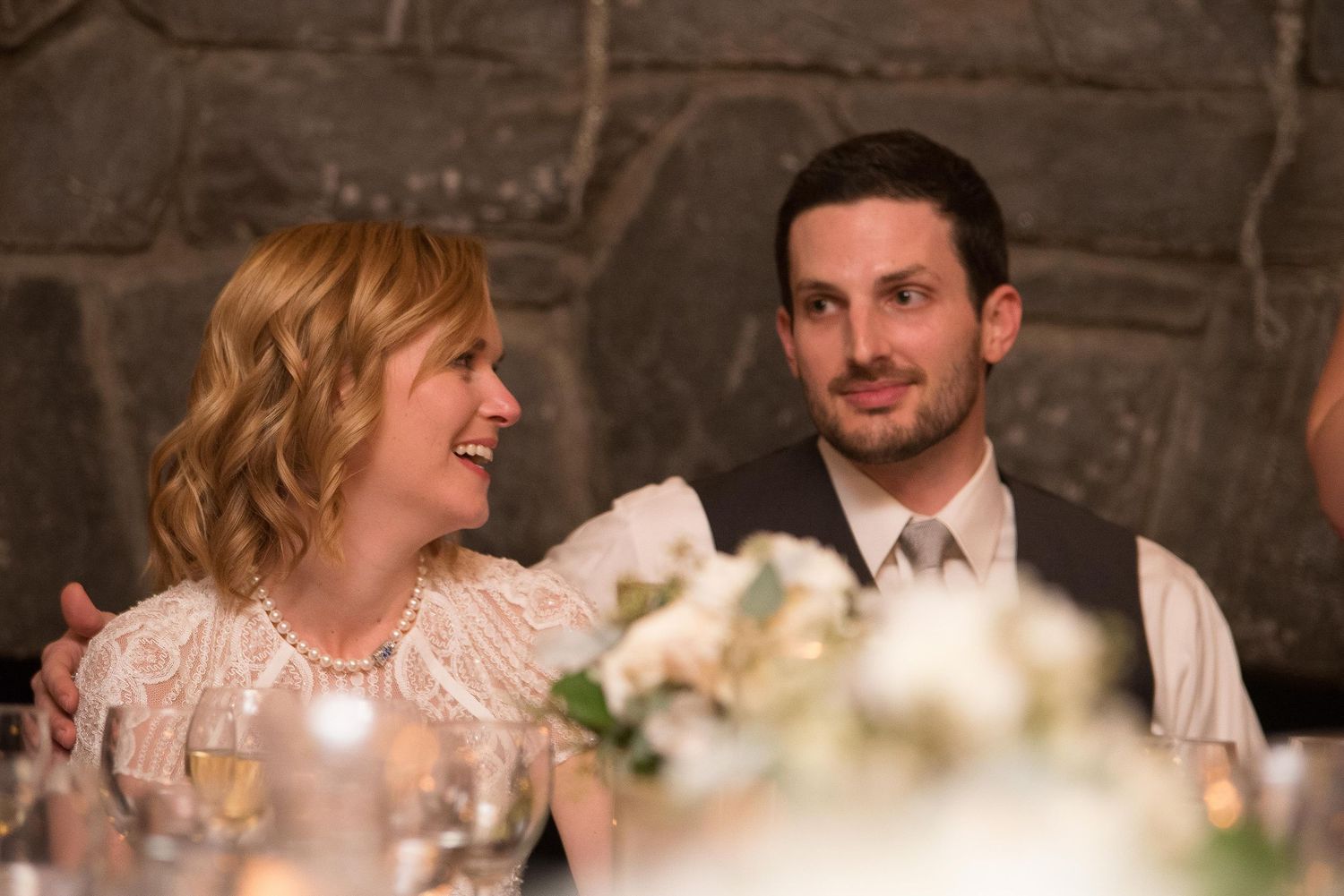 Couple smiling and laughing together at a wedding reception dinner, woman in white lace dress with pearl necklace.