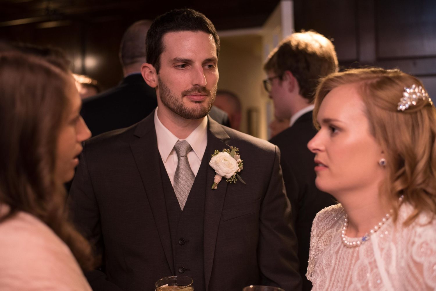 A groom in a gray suit with boutonniere converses with two women at a wedding reception.