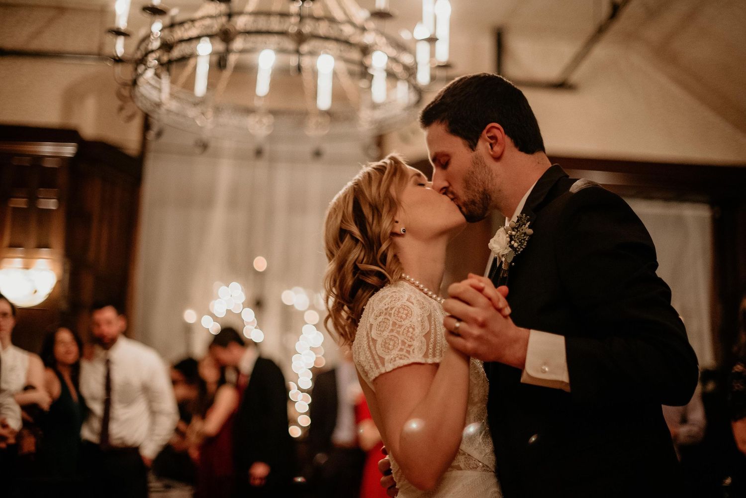 Bride and groom share first dance kiss under chandelier at elegant wedding reception.