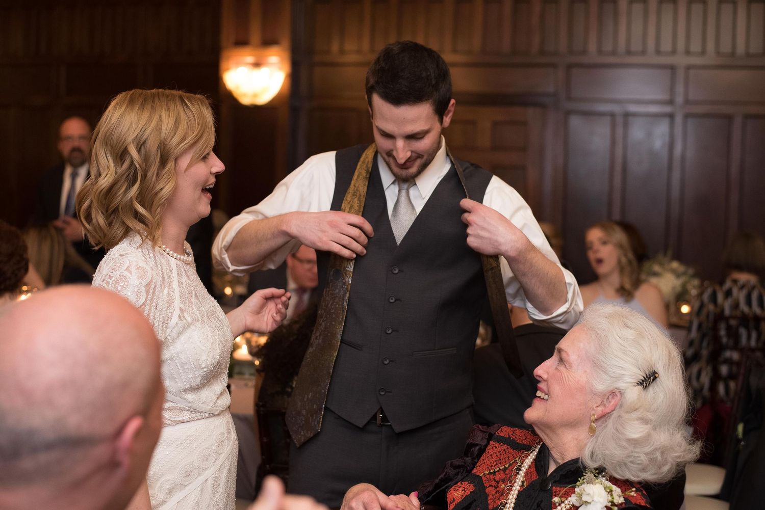 Groom in vest laughing with bride and elderly guest at elegant wood-paneled wedding reception.
