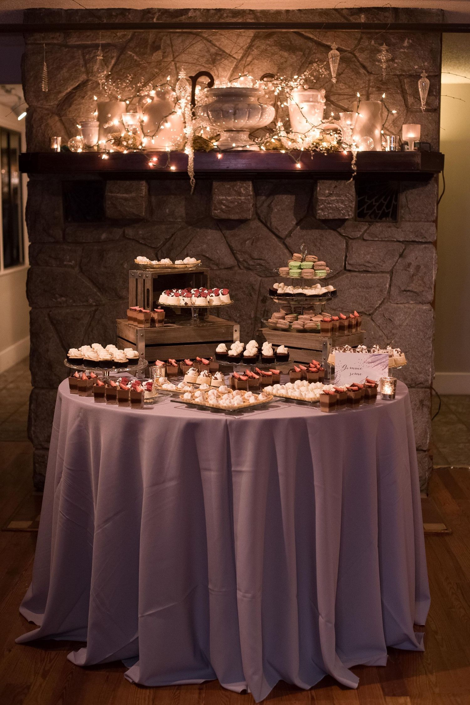 Elegant dessert table with lavender tablecloth in front of a stone fireplace adorned with warm string lights.