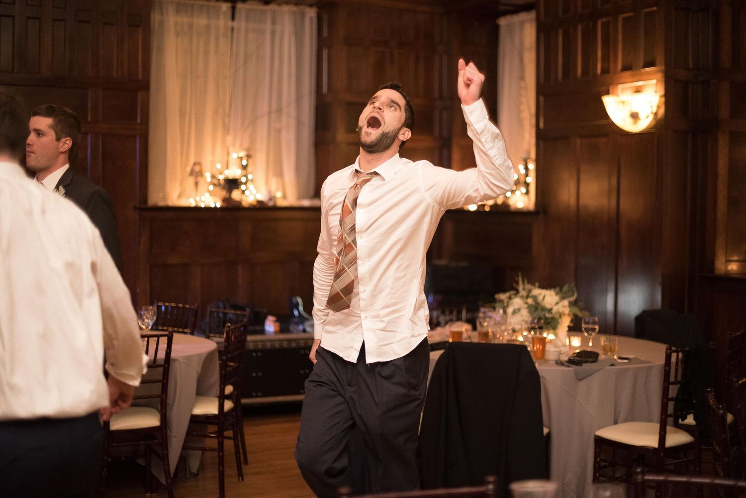 Man in white shirt and gold tie dancing energetically at an elegant wood-paneled wedding reception venue.