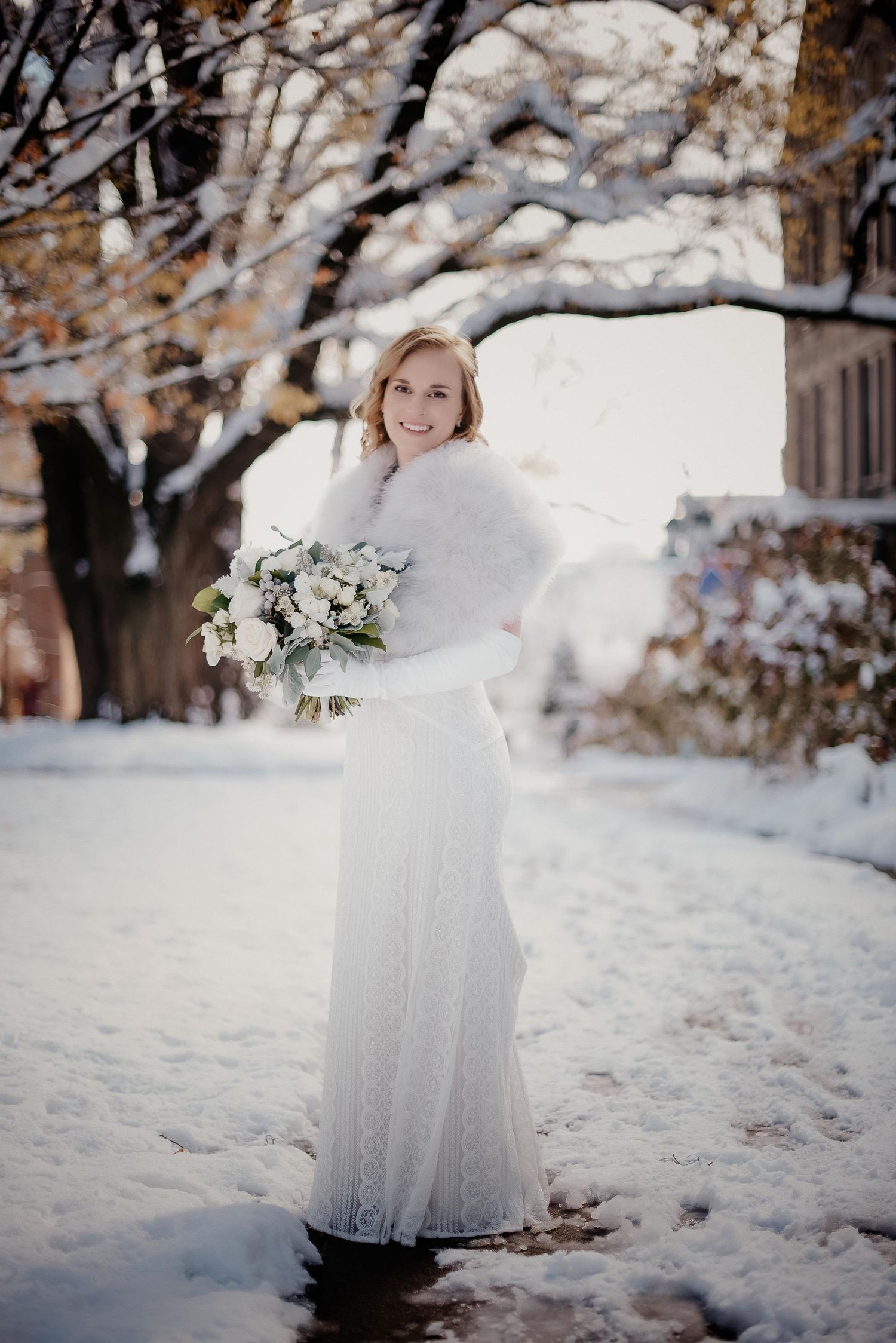 Bride in white lace gown and fur wrap holding bouquet, posing in snowy winter landscape with snow-covered trees.