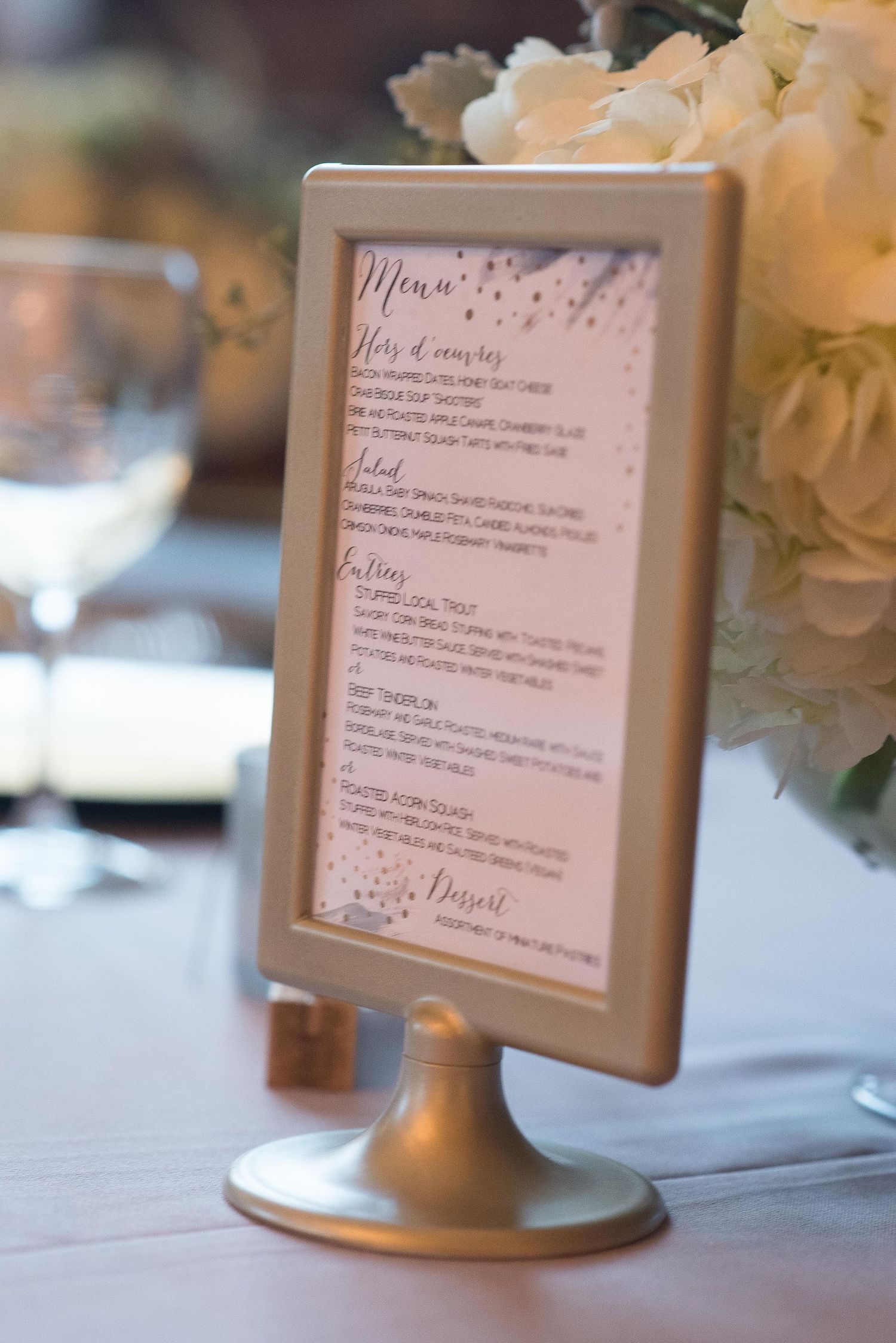 Elegant gold framed wedding menu card on a pedestal stand with floral centerpiece at a reception table.