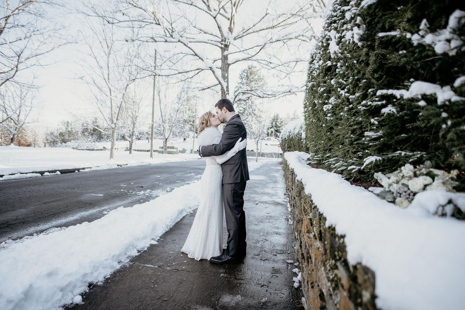 A bride and groom share a romantic embrace outdoors in a snowy winter wedding setting beside a stone wall.