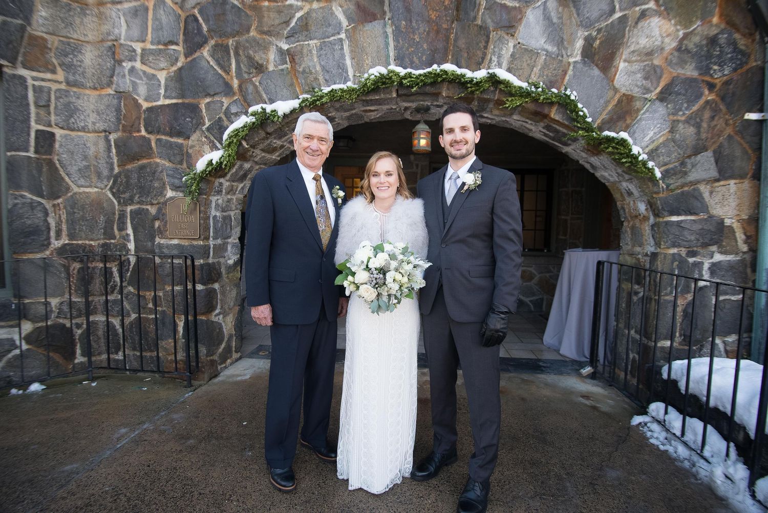 Bride in white gown stands with two men in suits before a stone archway decorated with greenery and snow.
