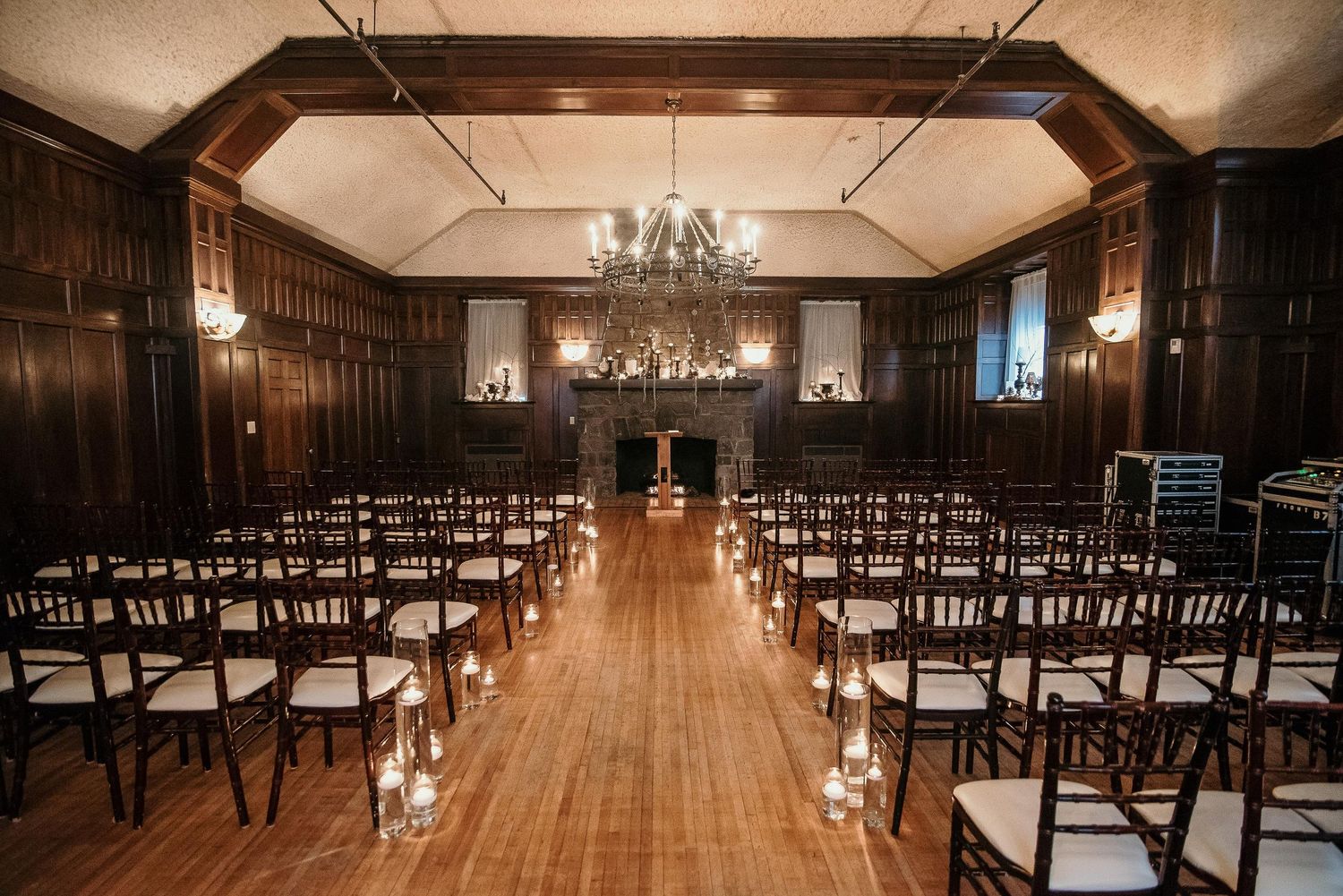 Elegant wedding ceremony setup in dark wood-paneled room with chandelier, chiavari chairs, and candlelit aisle.