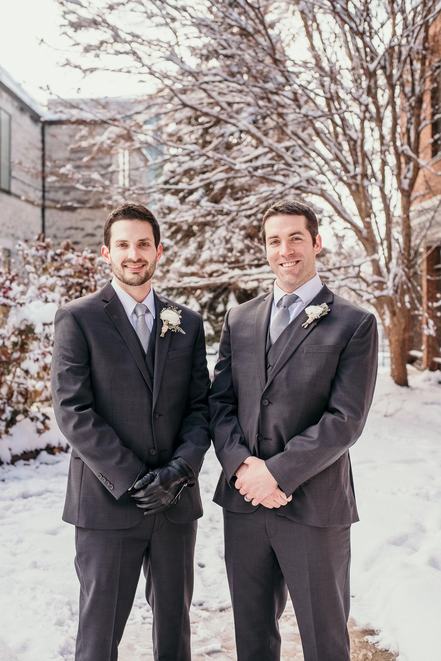 Two groomsmen in dark suits with boutonnieres pose outdoors in a snowy winter wedding setting.