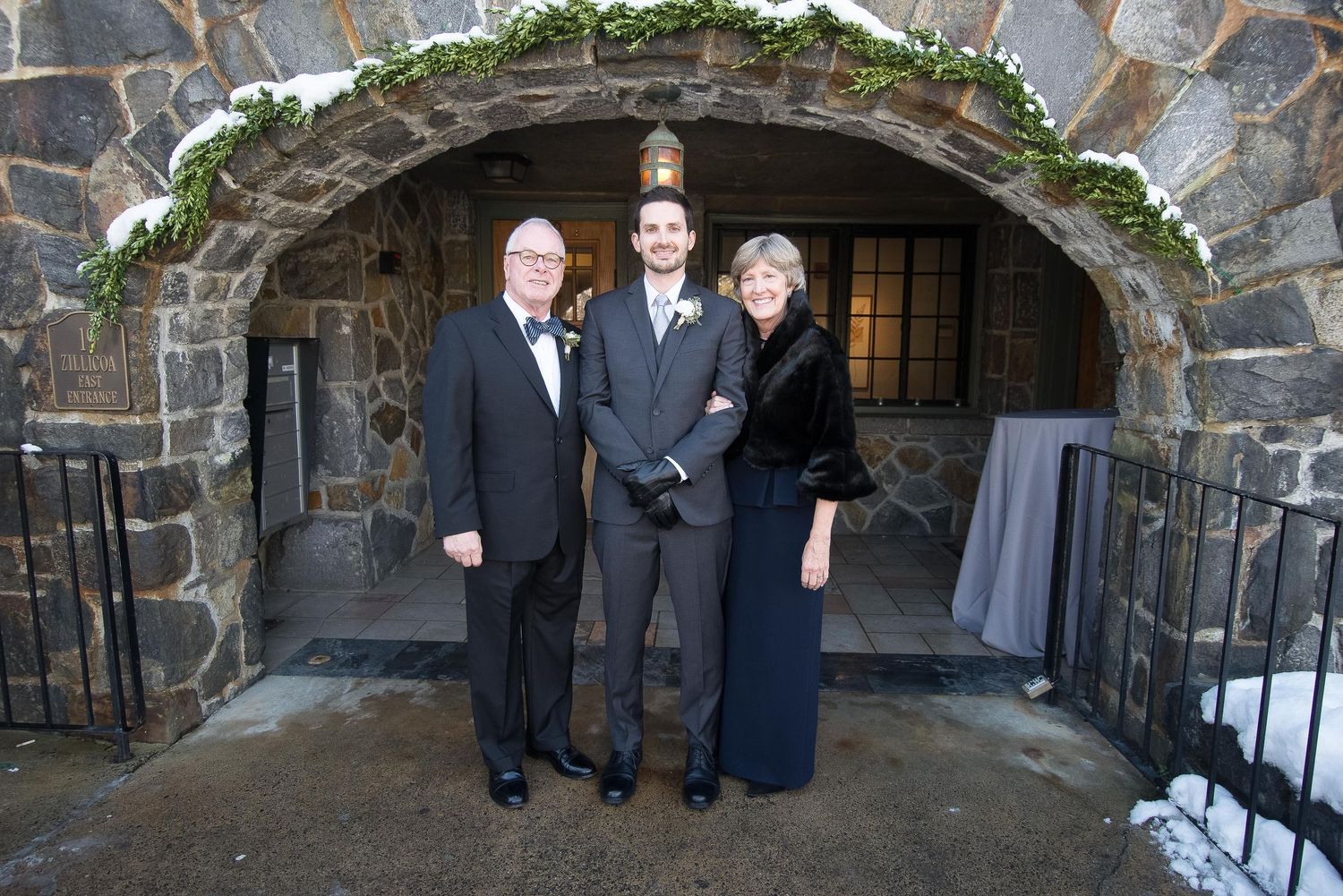 Three people posing under a stone archway at a winter wedding venue, dressed in formal attire.