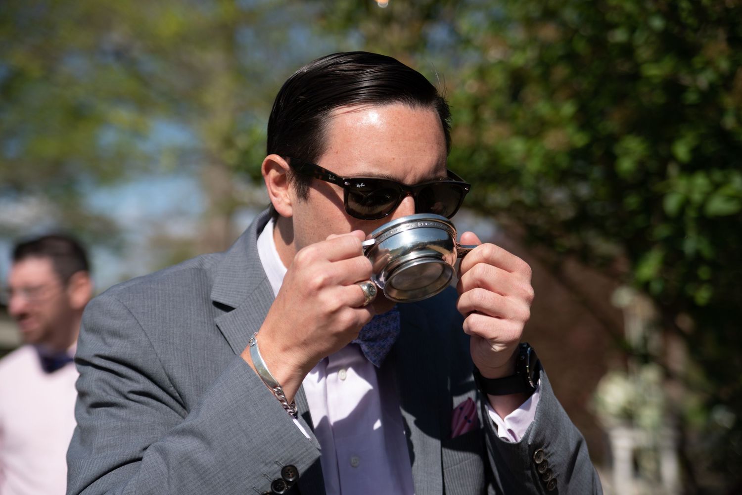 A wedding guest in a gray suit drinks from a silver cup at an outdoor wedding celebration.