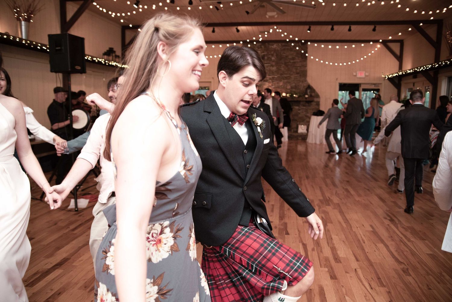 Couple dancing energetically at a wedding reception, man wearing a red tartan kilt and bow tie in a rustic barn venue.