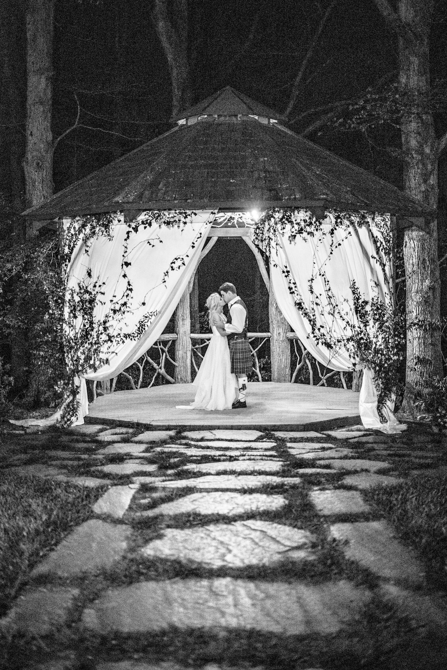 Black and white photo of a couple sharing a kiss under a decorated gazebo with draped fabric and stone pathway at night.