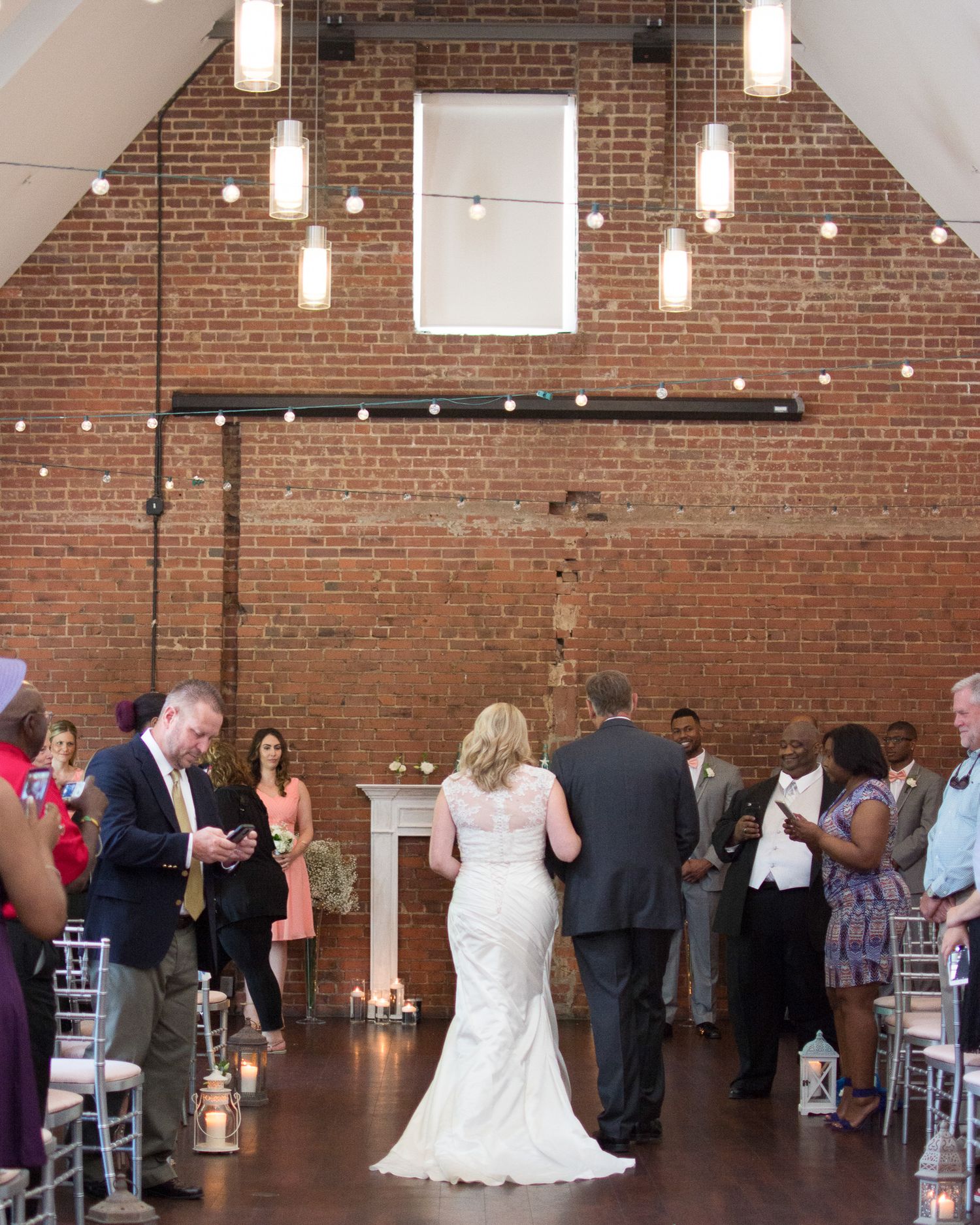 Bride walking down the aisle in a lace wedding gown inside a rustic brick venue with hanging Edison bulb lights.