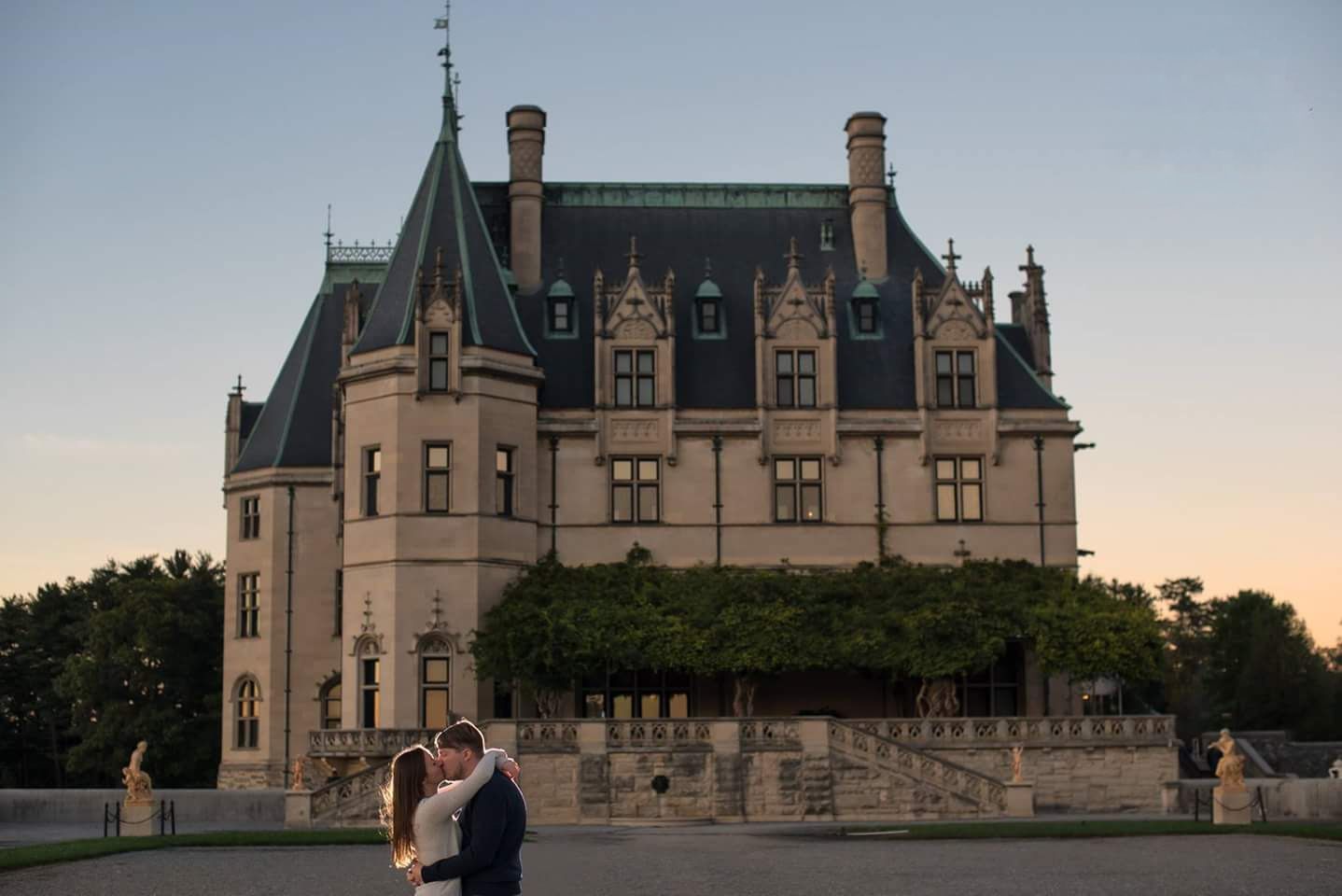Couple sharing a romantic embrace in front of the grand Biltmore Estate at dusk.