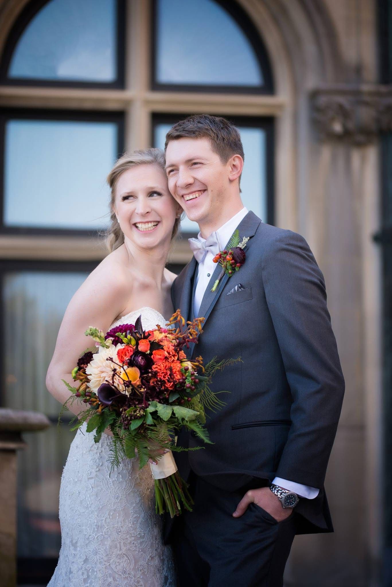 Smiling bride and groom posing outdoors with a vibrant autumn bouquet of orange and red flowers.