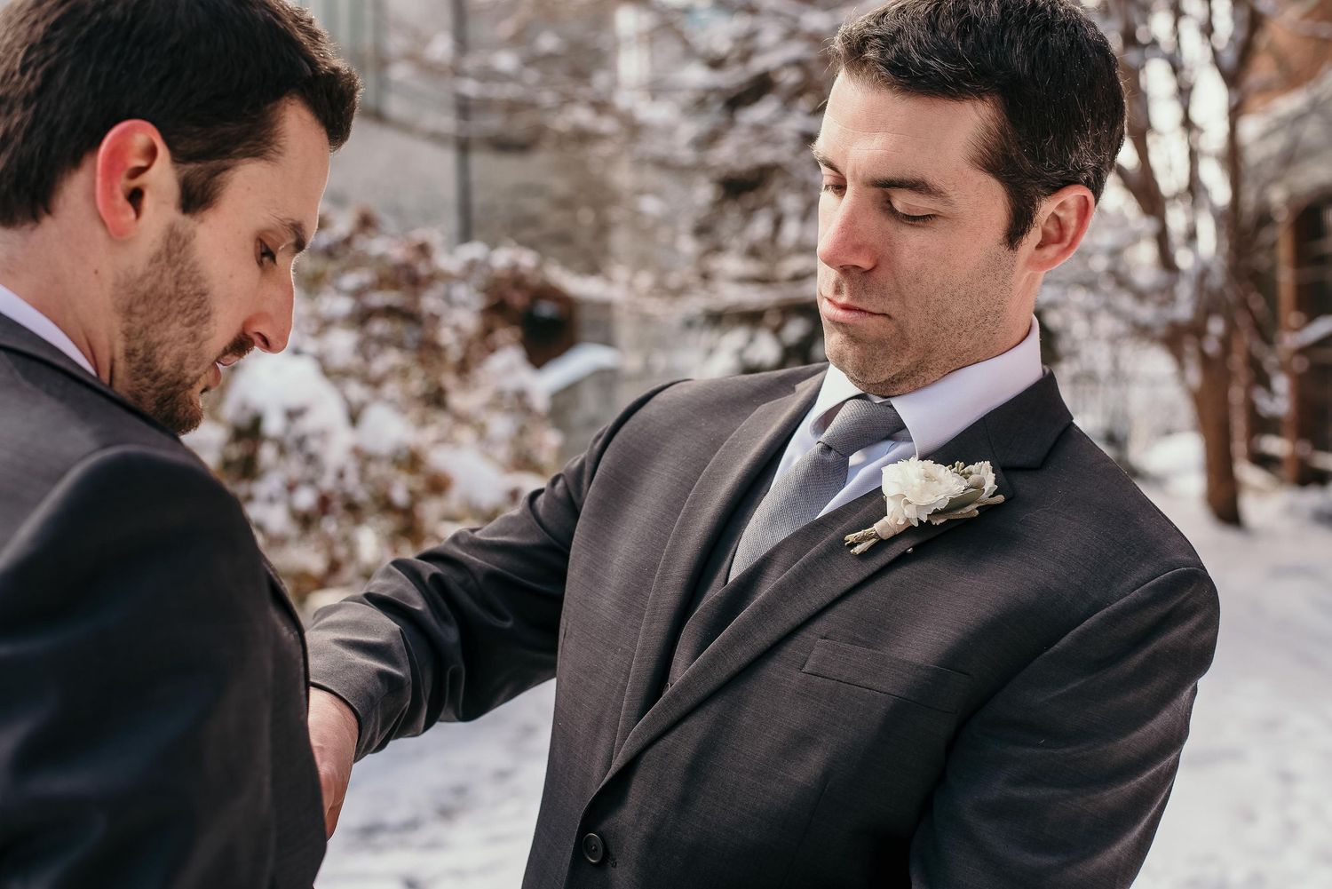 Two men in gray suits adjusting boutonniere outdoors in snowy winter wedding setting.
