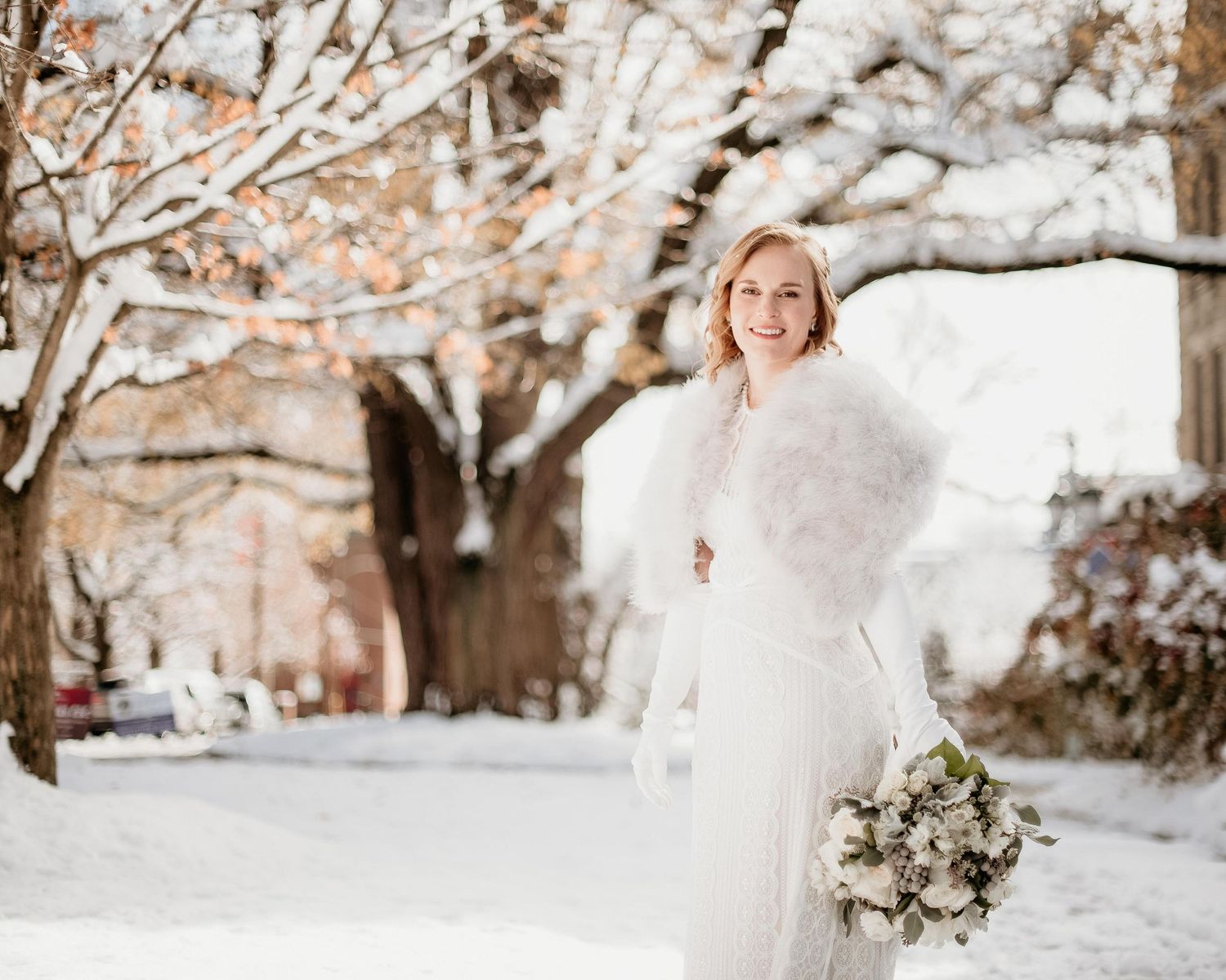 Smiling bride in white gown and fur stole holding bouquet in snowy winter wonderland setting.