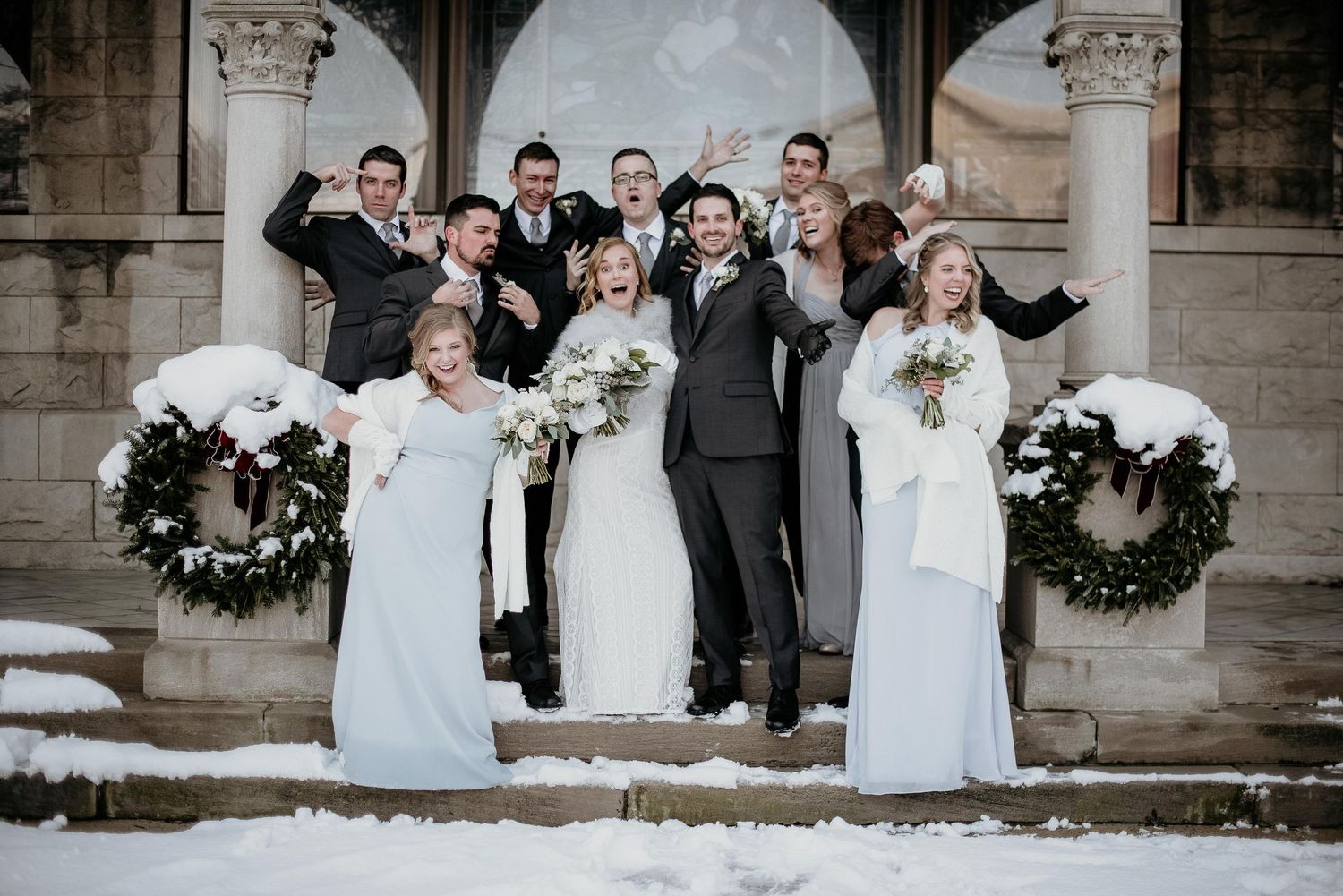 Winter wedding party poses on snowy steps, bride and groom centered with bridesmaids and groomsmen celebrating outdoors.