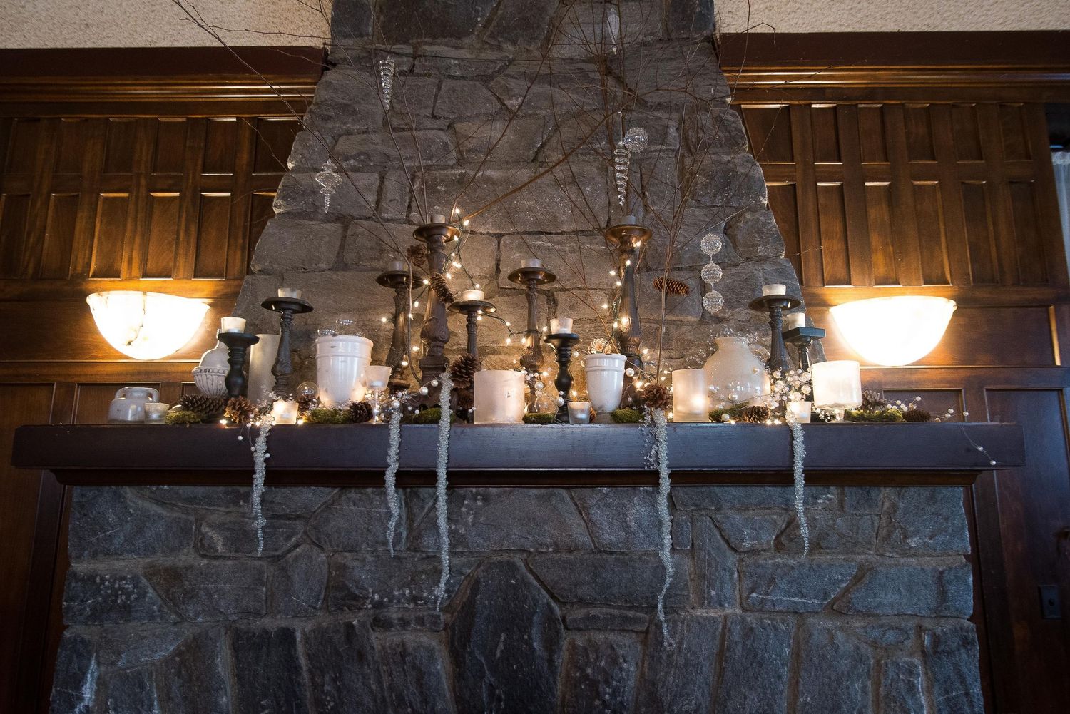 Stone fireplace mantel decorated with white candles, fairy lights, and icicle garland for a winter holiday display.