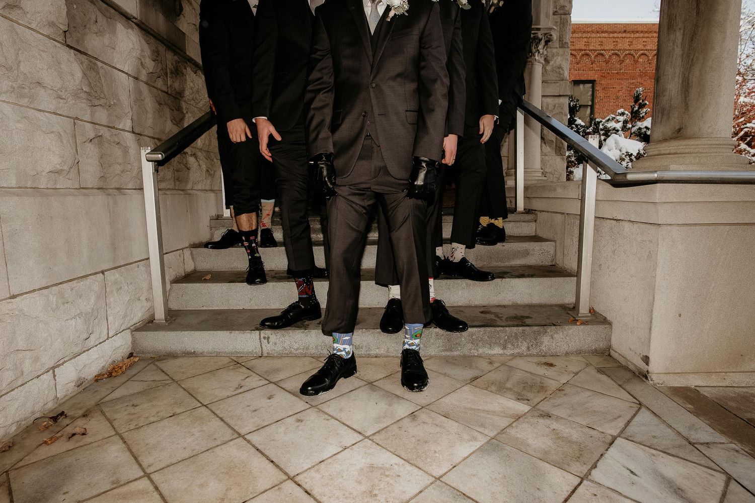 Groomsmen in dark suits displaying fun colorful socks on stone steps during a wedding celebration.