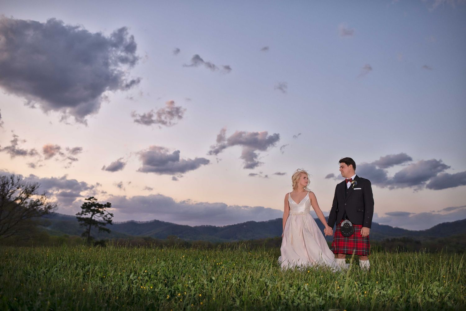 Bride and groom in Scottish kilt holding hands in green meadow at sunset with mountain backdrop.