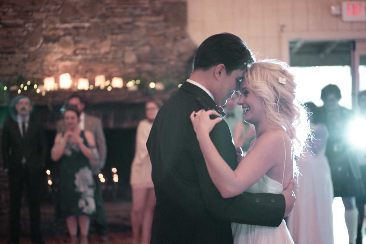 Bride and groom share their first dance at a rustic wedding reception with guests watching and photographing.