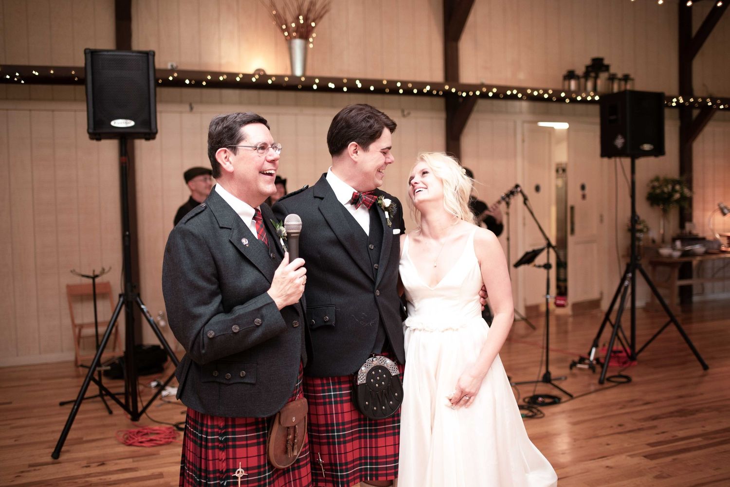 Wedding reception toast with groom in tuxedo, bride in white gown, and officiant in red tartan kilt.