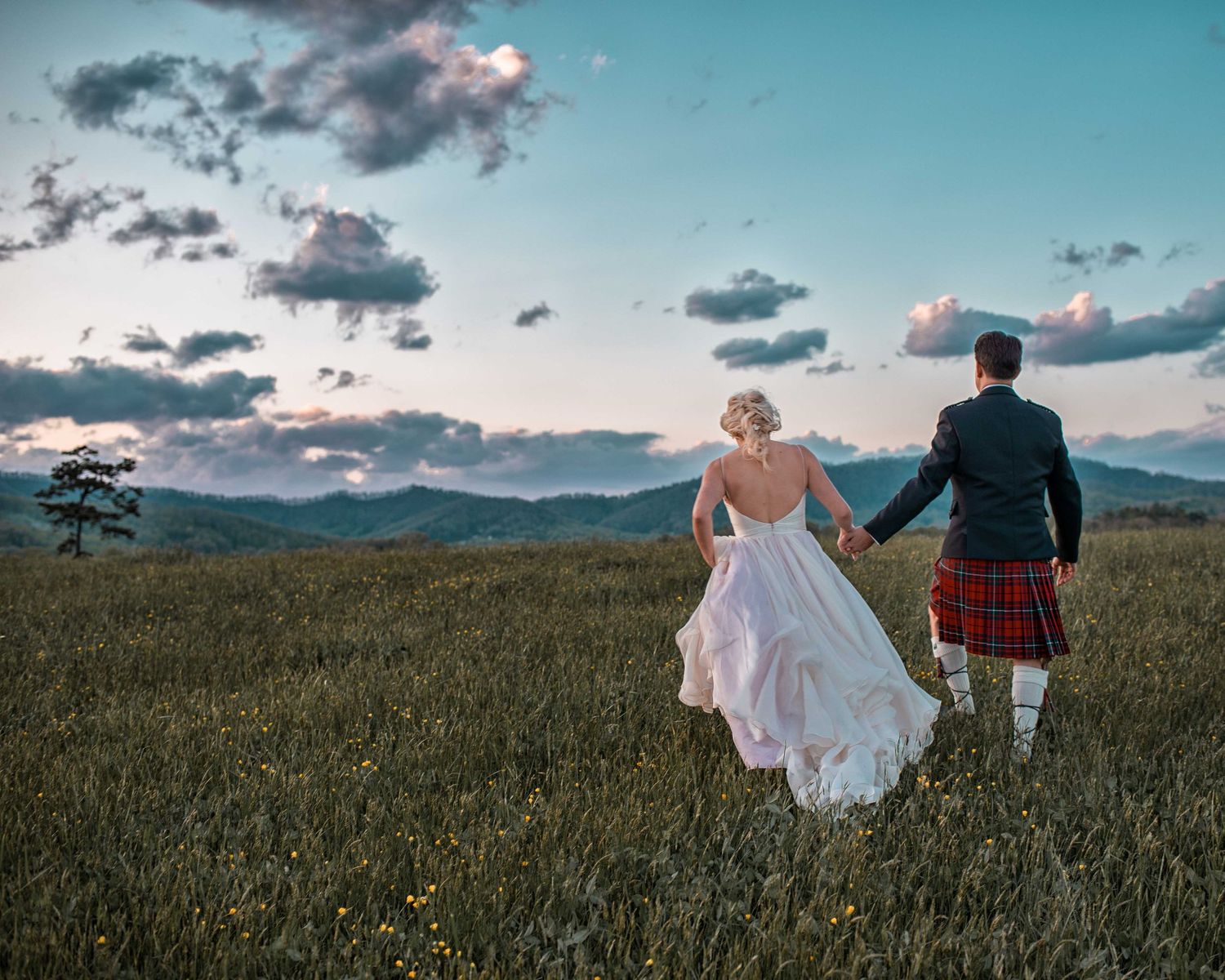 Bride and groom in Scottish kilt holding hands walking through green meadow with mountain sunset backdrop.