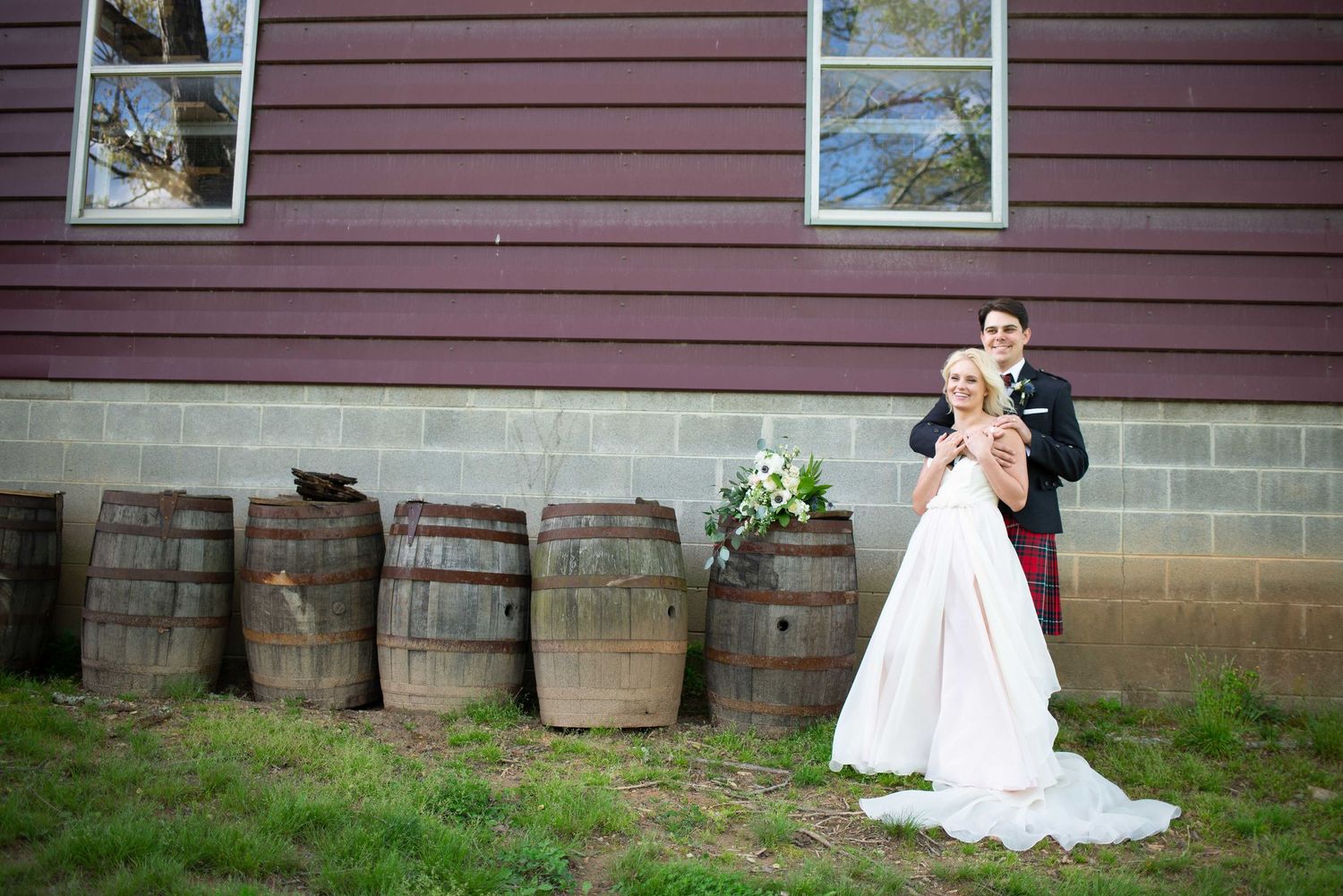 Bride and groom posing beside rustic wooden barrels outside a red barn at a country wedding venue.