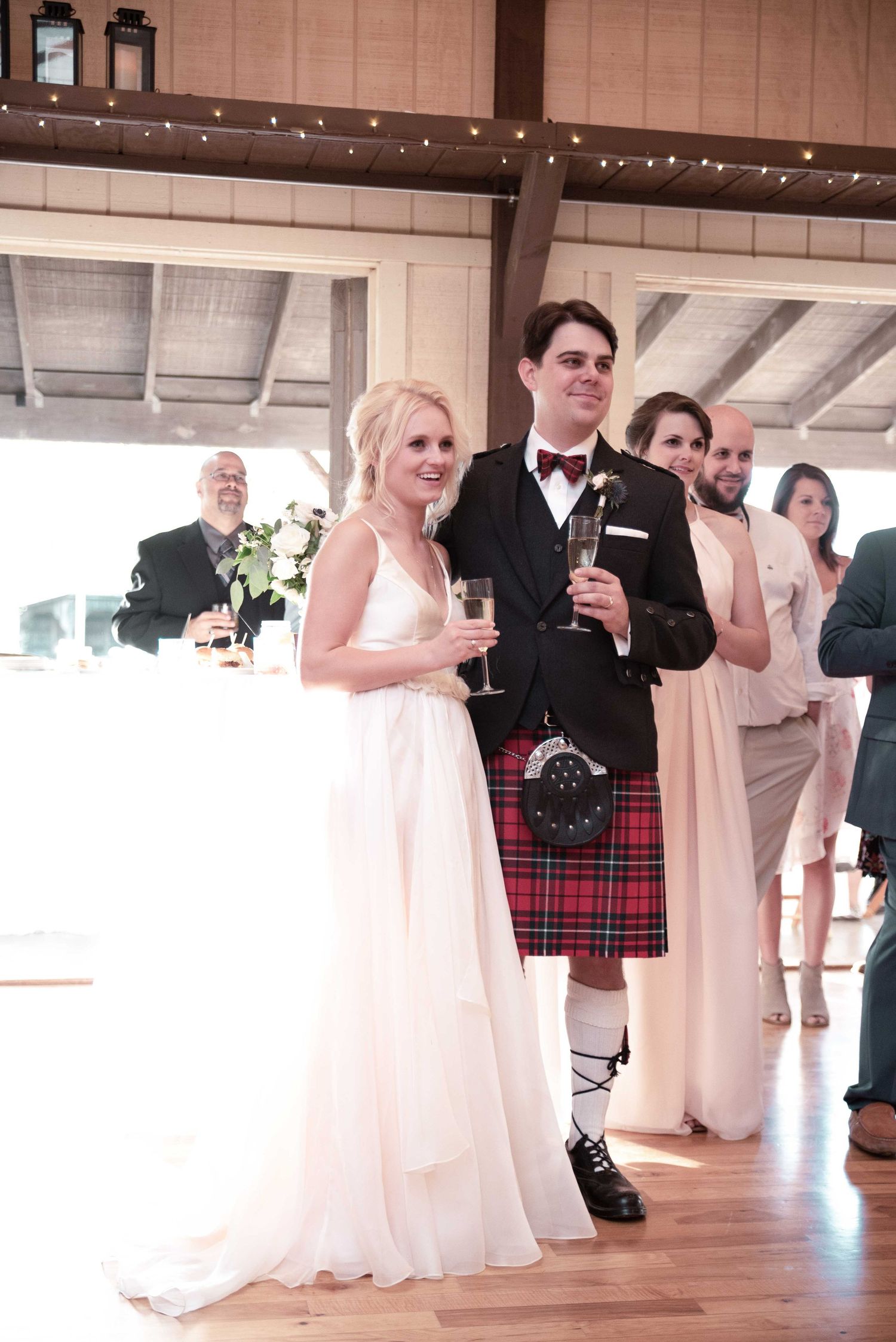 Bride in white gown and groom in red tartan kilt toast at wedding reception with guests in background.