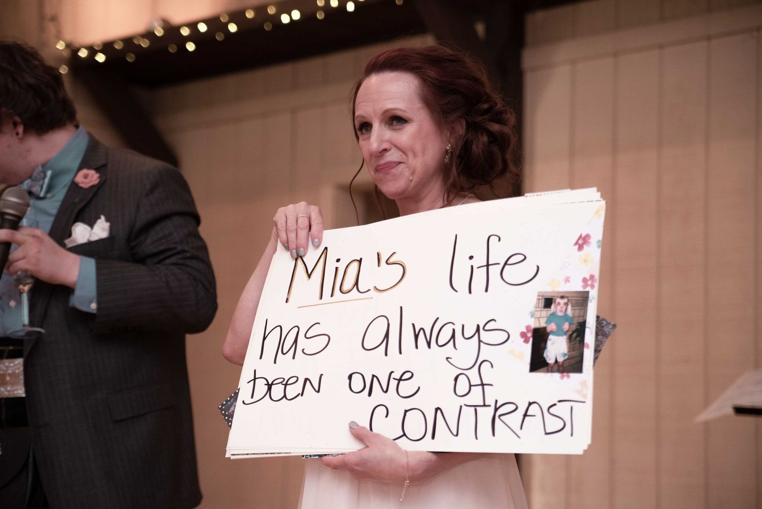 Woman holding handmade sign reading Mia's life has always been one of CONTRAST at a wedding reception.