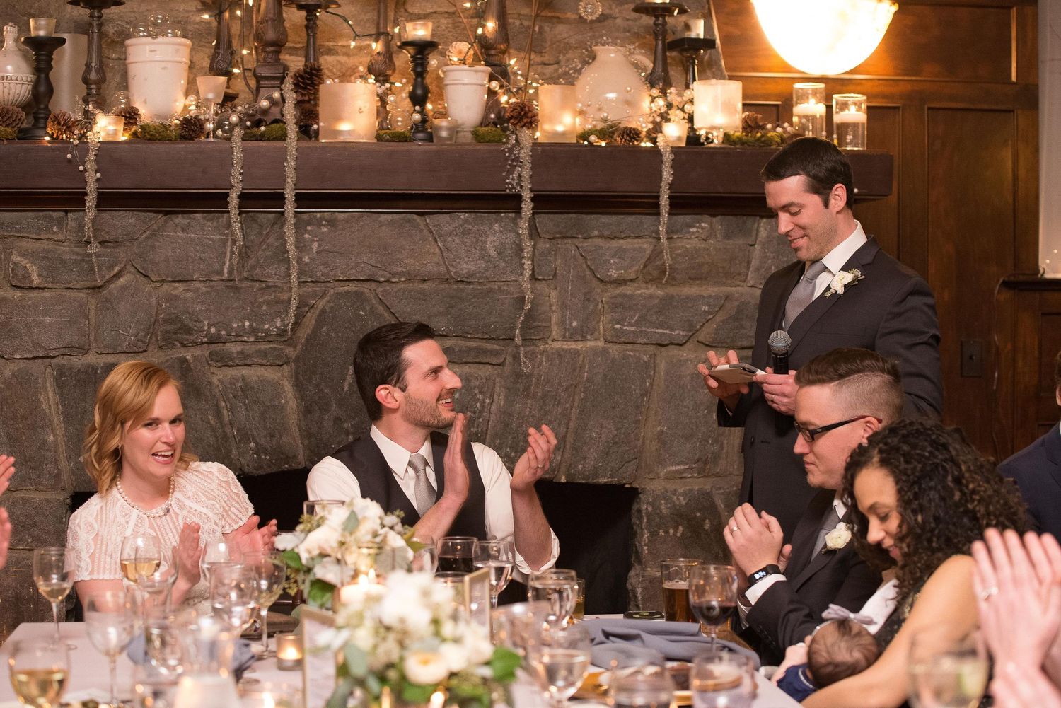 Wedding reception toast with guests applauding as groom delivers speech near stone fireplace.