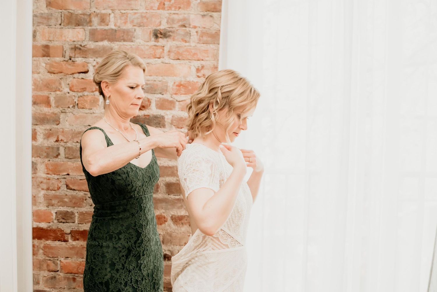 A bride in a white gown gets help fastening her dress from a woman in a green lace dress near a brick wall.