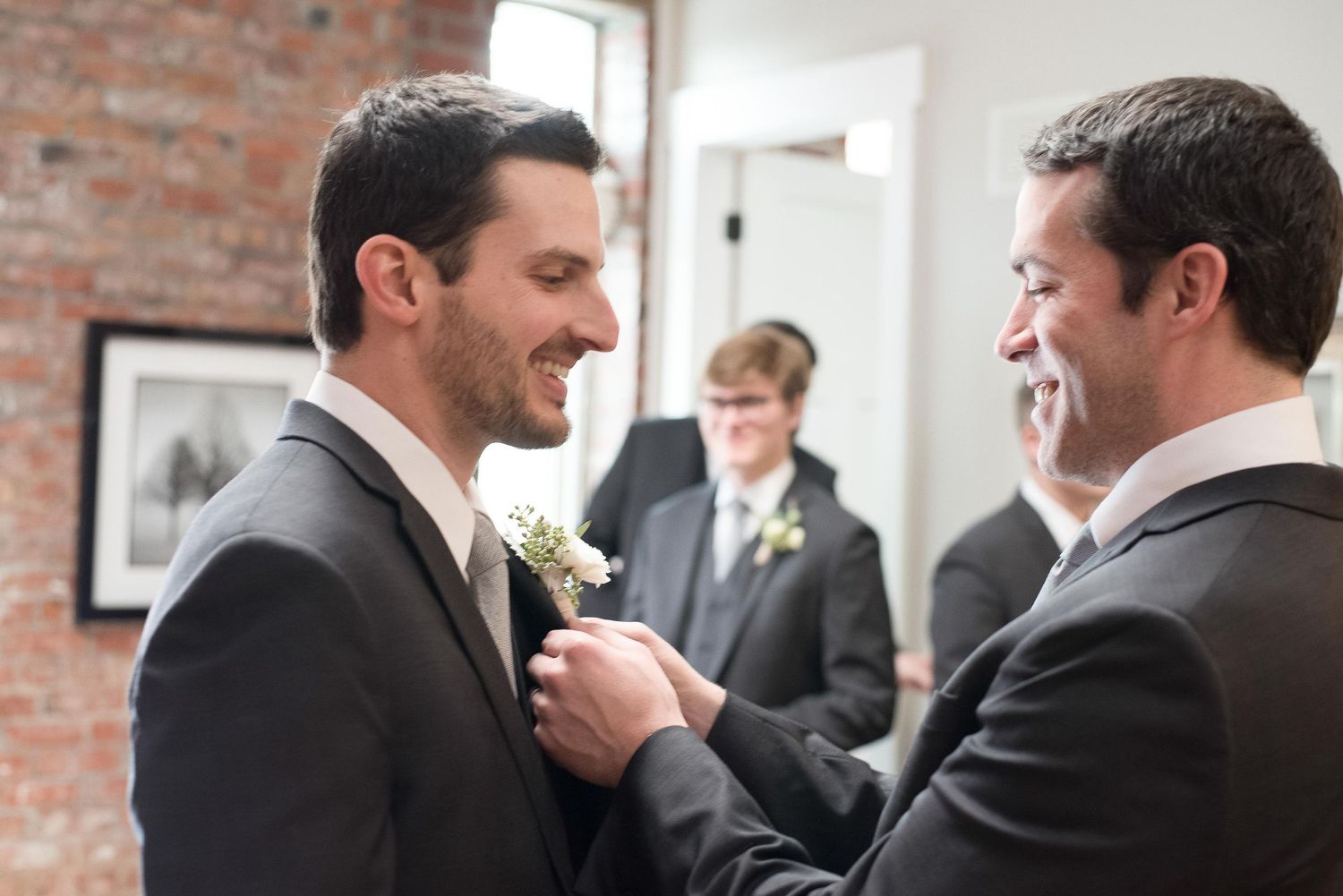 Groomsman pinning a boutonniere on the groom's lapel before a wedding ceremony in a rustic venue.