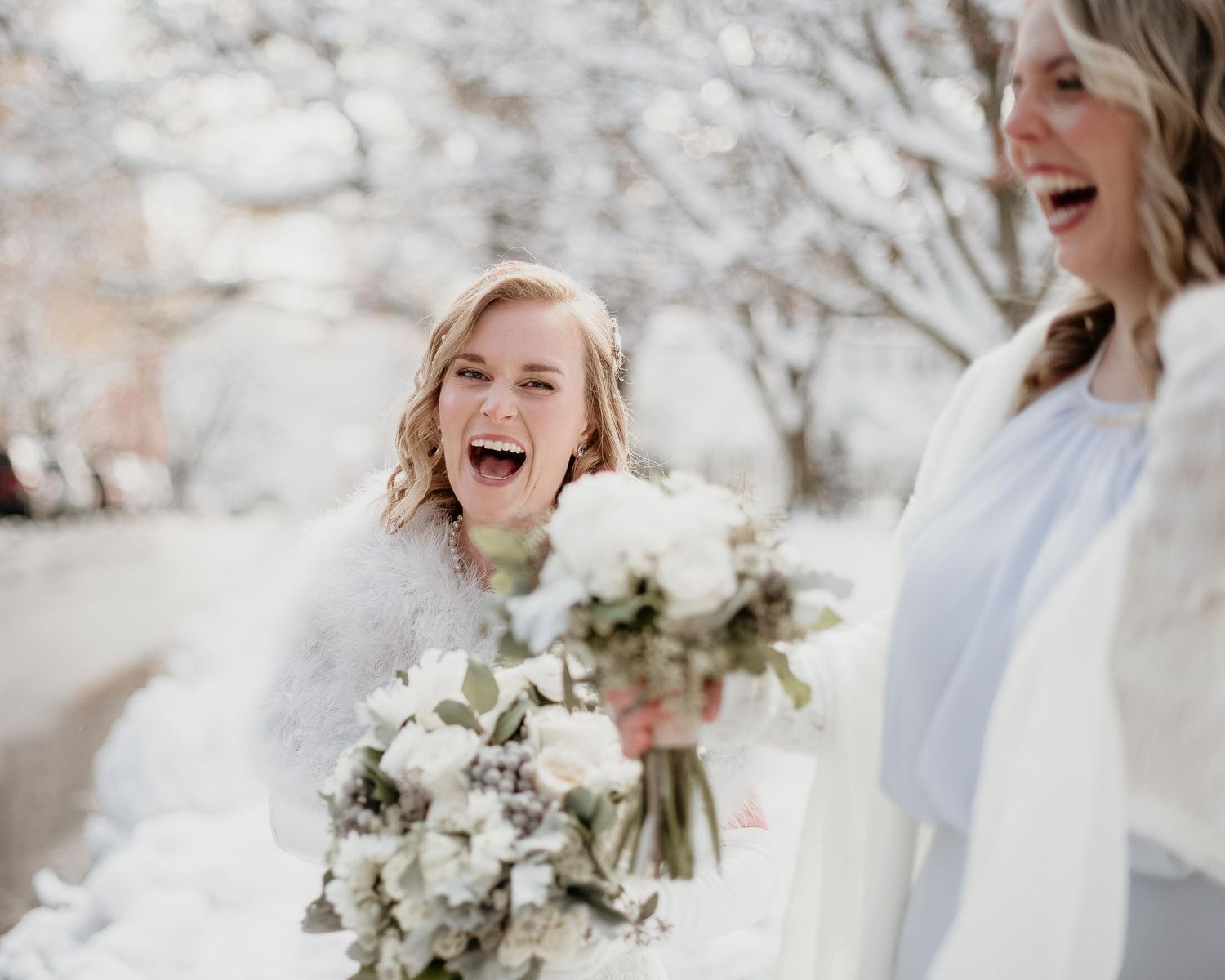 Two joyful brides laughing in snow holding white floral bouquets in a winter wedding setting.