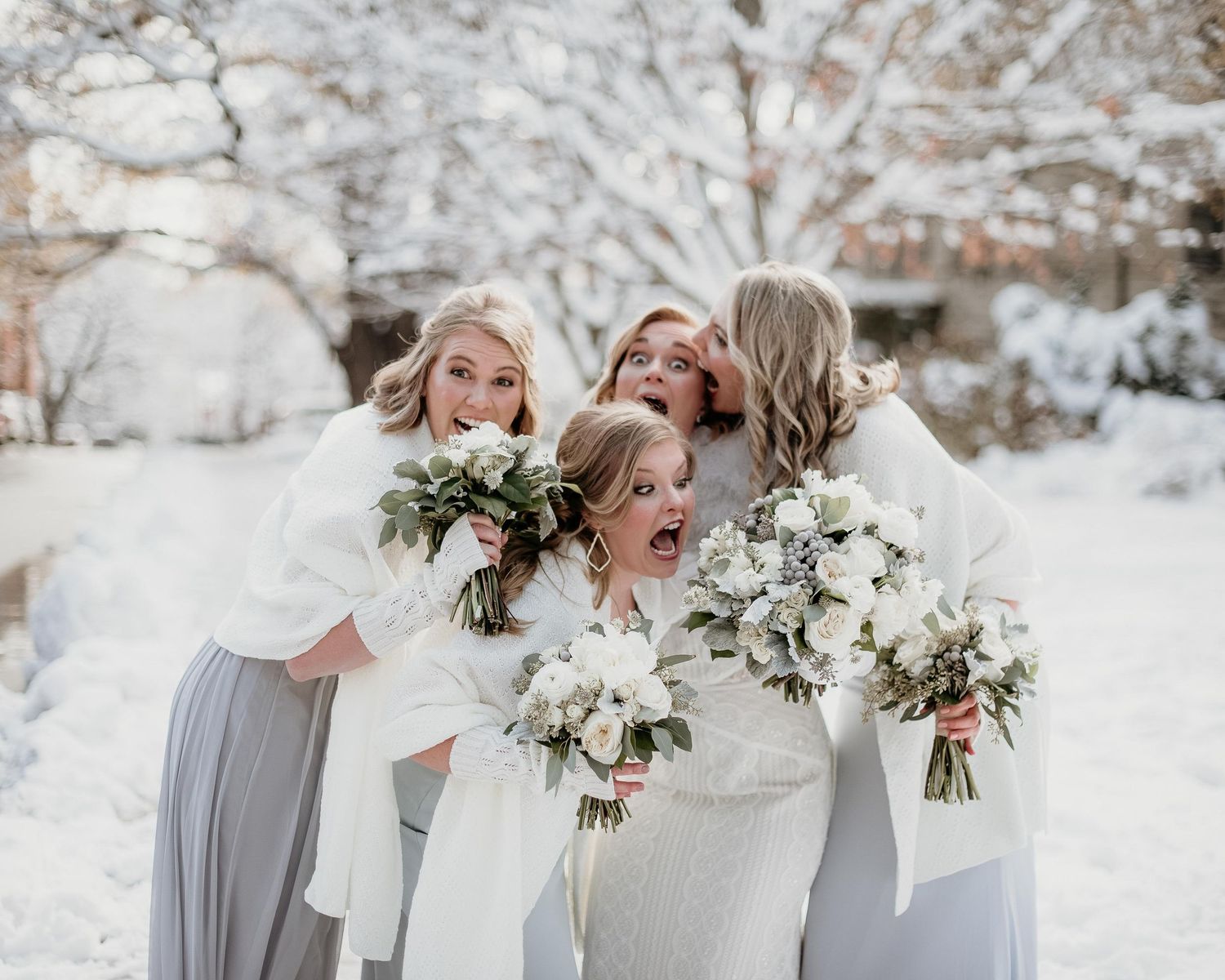 Bride and bridesmaids in white dresses holding bouquets laughing together in a snowy winter wedding outdoor setting.
