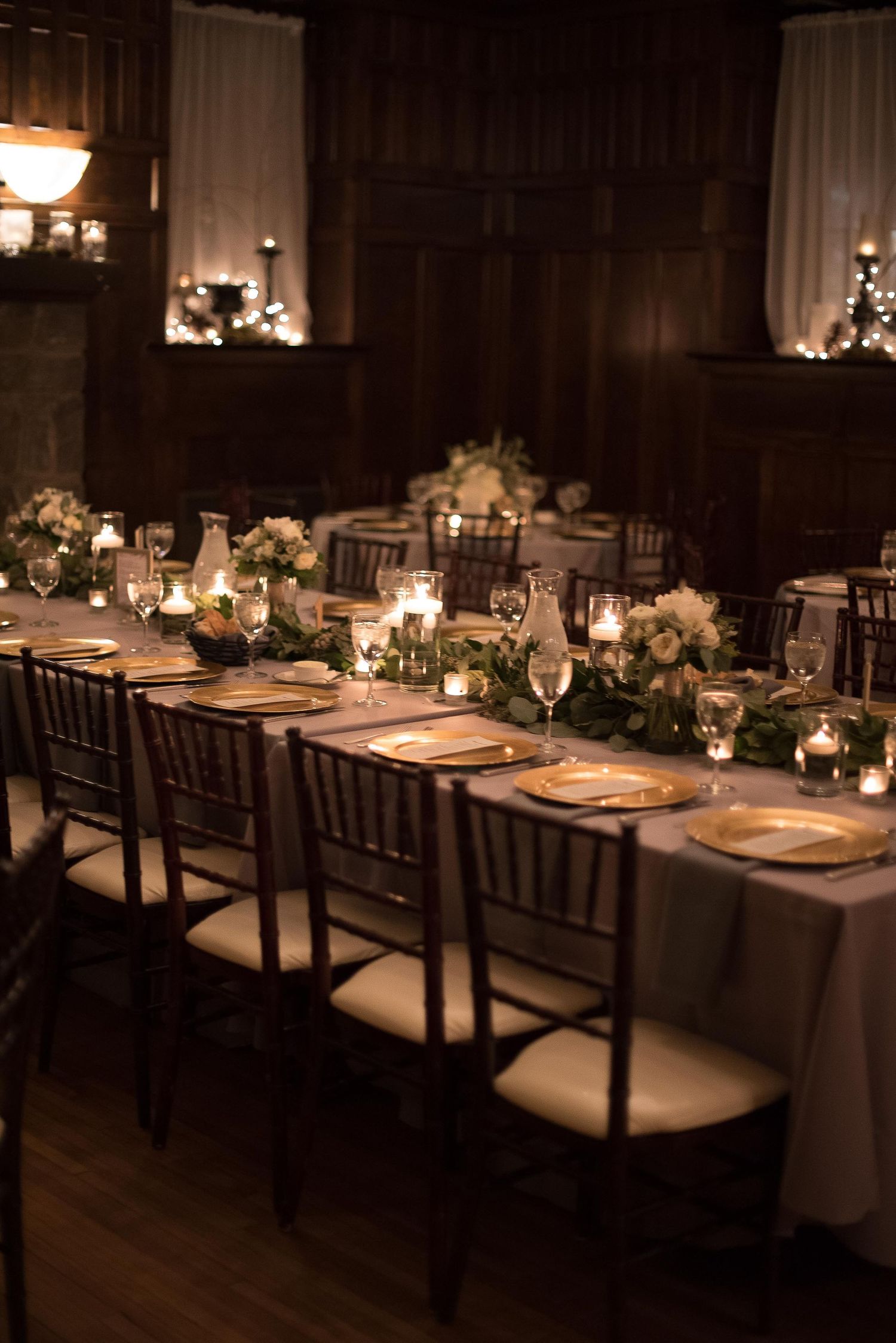 Elegant candlelit wedding reception with long tables, gold chargers, floral garlands, and dark wood Chiavari chairs.