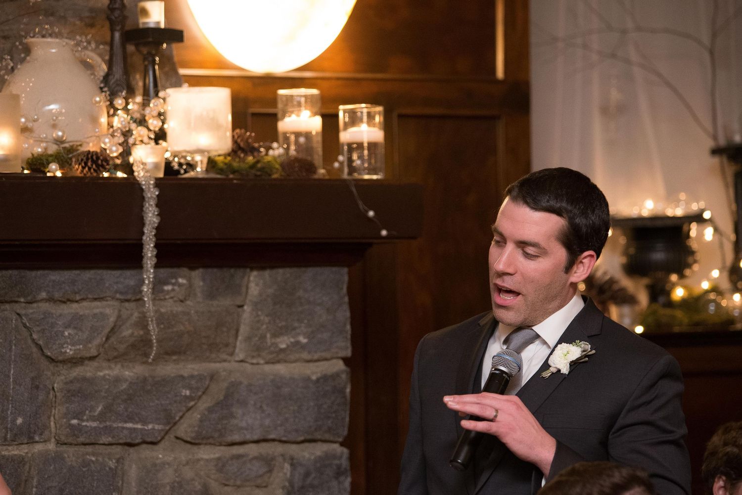 Man in suit giving speech at elegant winter wedding reception near fireplace with candles and holiday decor.