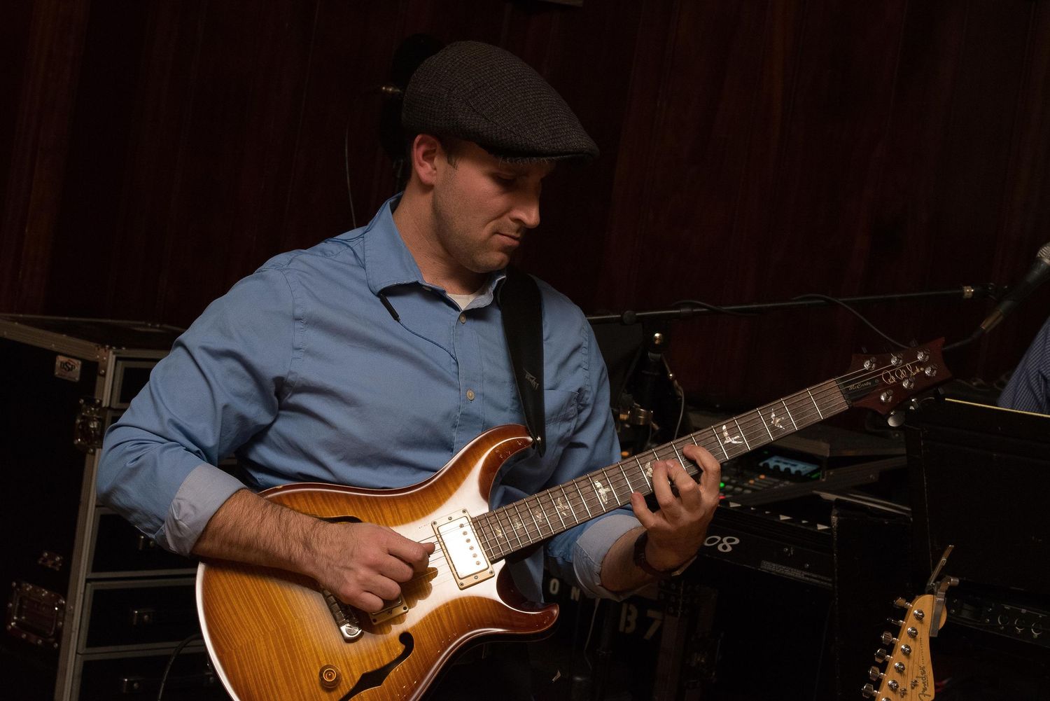 Musician playing an electric guitar on stage, wearing a gray flat cap and blue shirt under dim lighting.