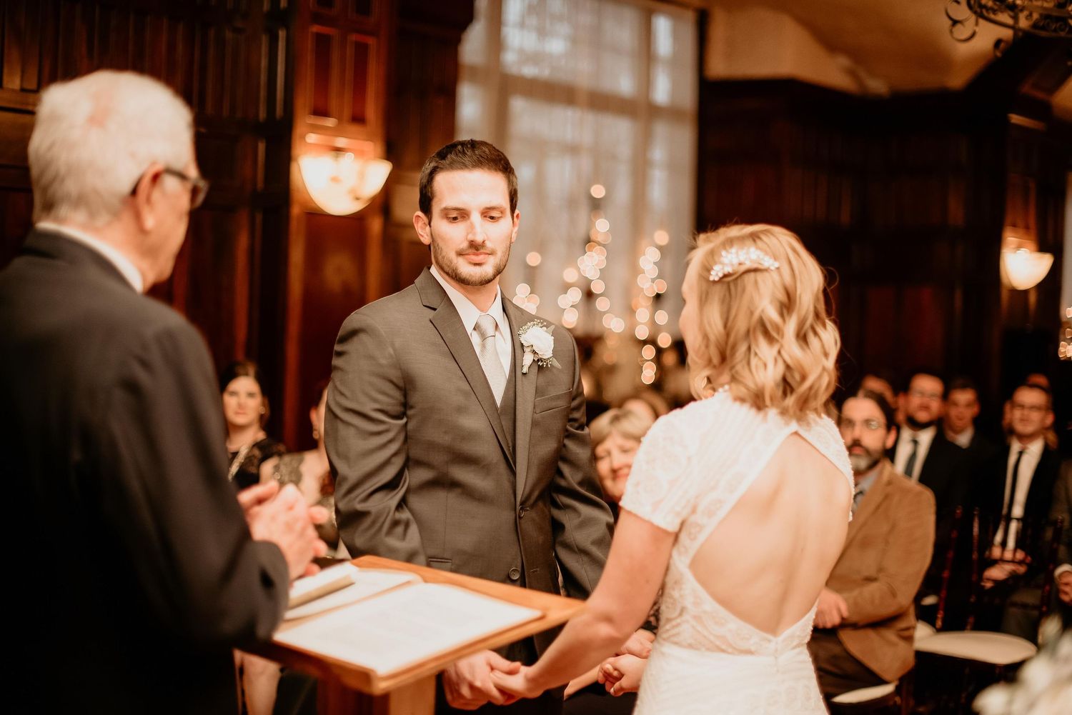 Groom holds bride's hands during indoor wedding ceremony in elegant wood-paneled venue with guests seated behind.