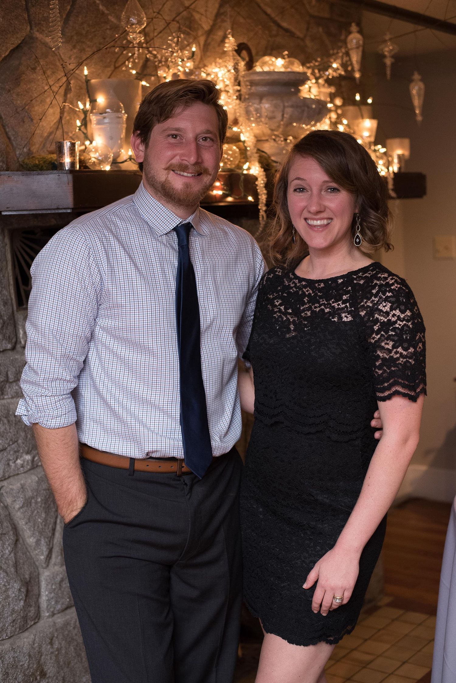 A smiling couple poses together in front of a decorated fireplace, man in dress shirt and tie, woman in black lace dress.