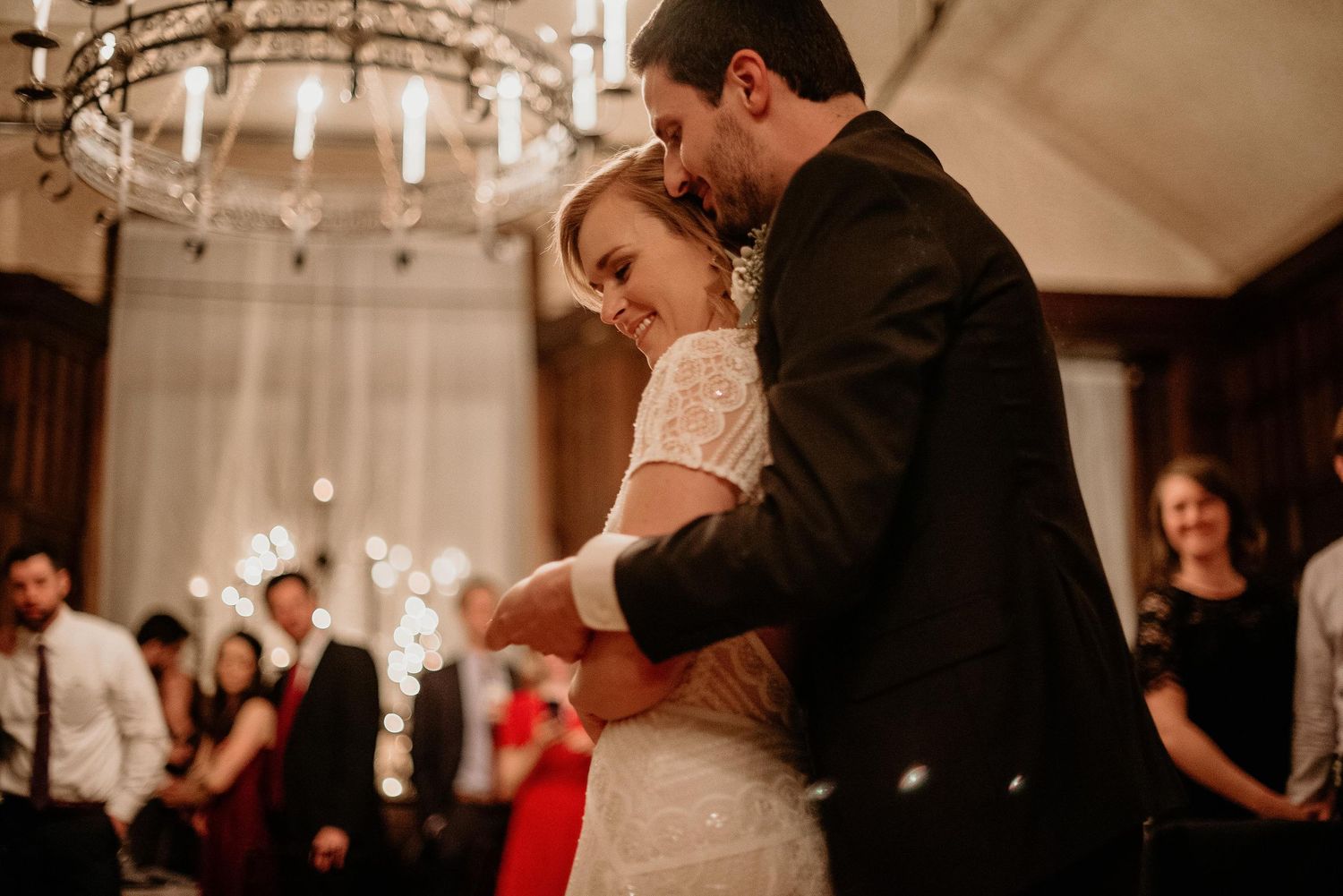 Bride and groom share their first dance under a chandelier while wedding guests watch and hold sparklers.