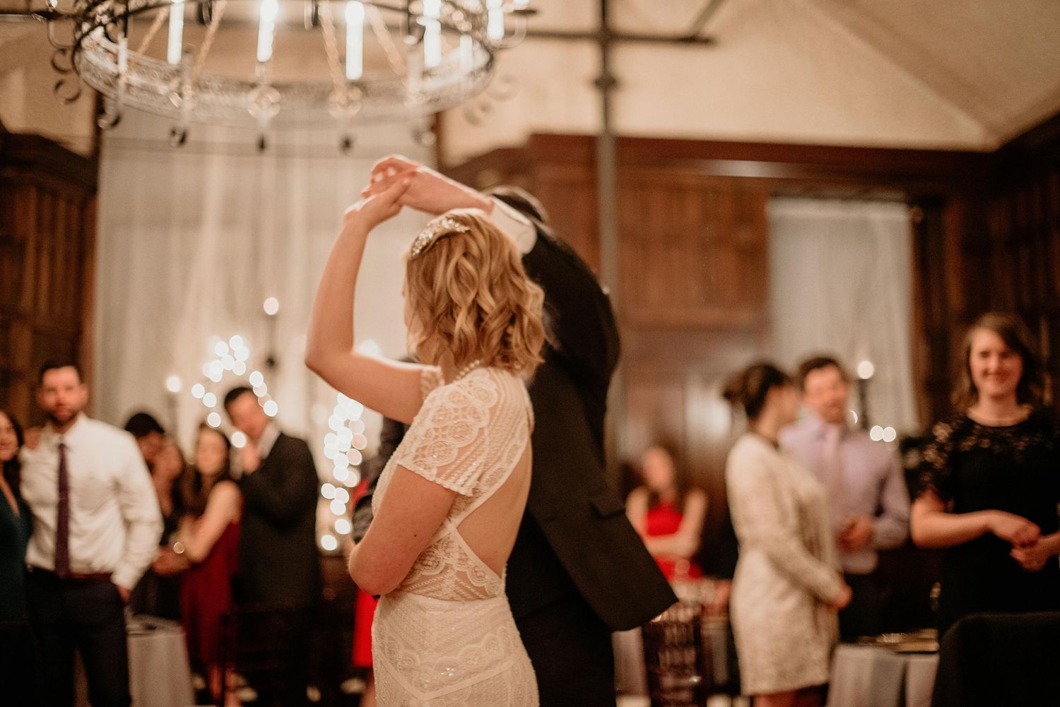 Bride and groom share first dance under chandelier at elegant wedding reception with guests watching.