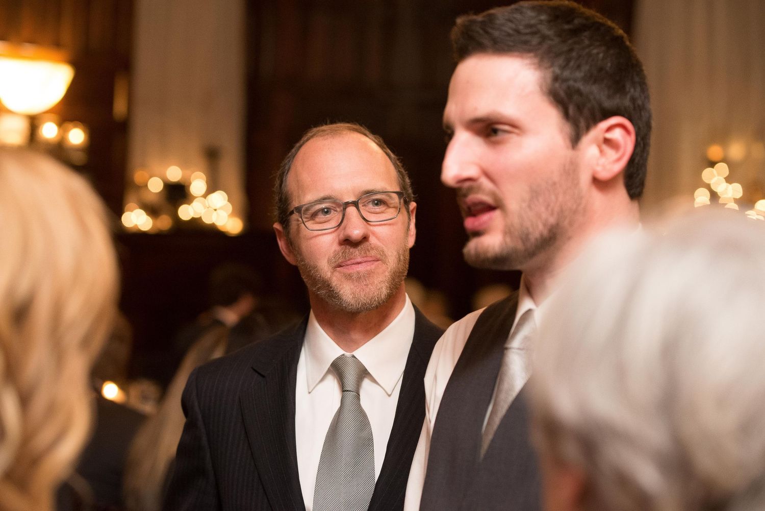 Two men in suits engage in conversation at an elegant formal event or wedding reception.