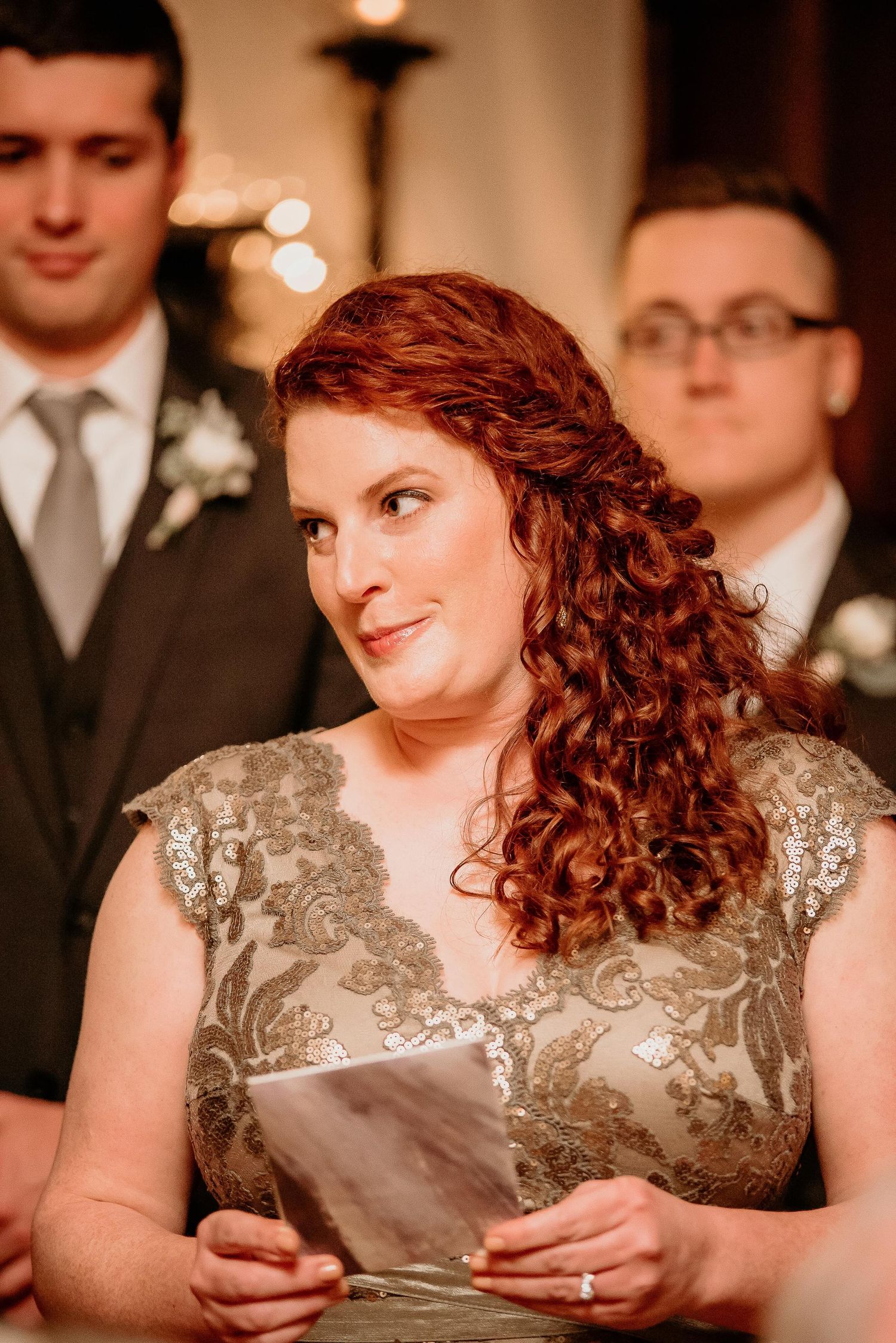 Red-haired woman in gold lace dress reads wedding vows at ceremony, with groomsmen standing behind her.
