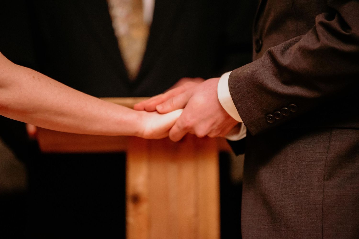 Couple holding hands during wedding ceremony, groom in brown suit, romantic close-up of clasped hands.